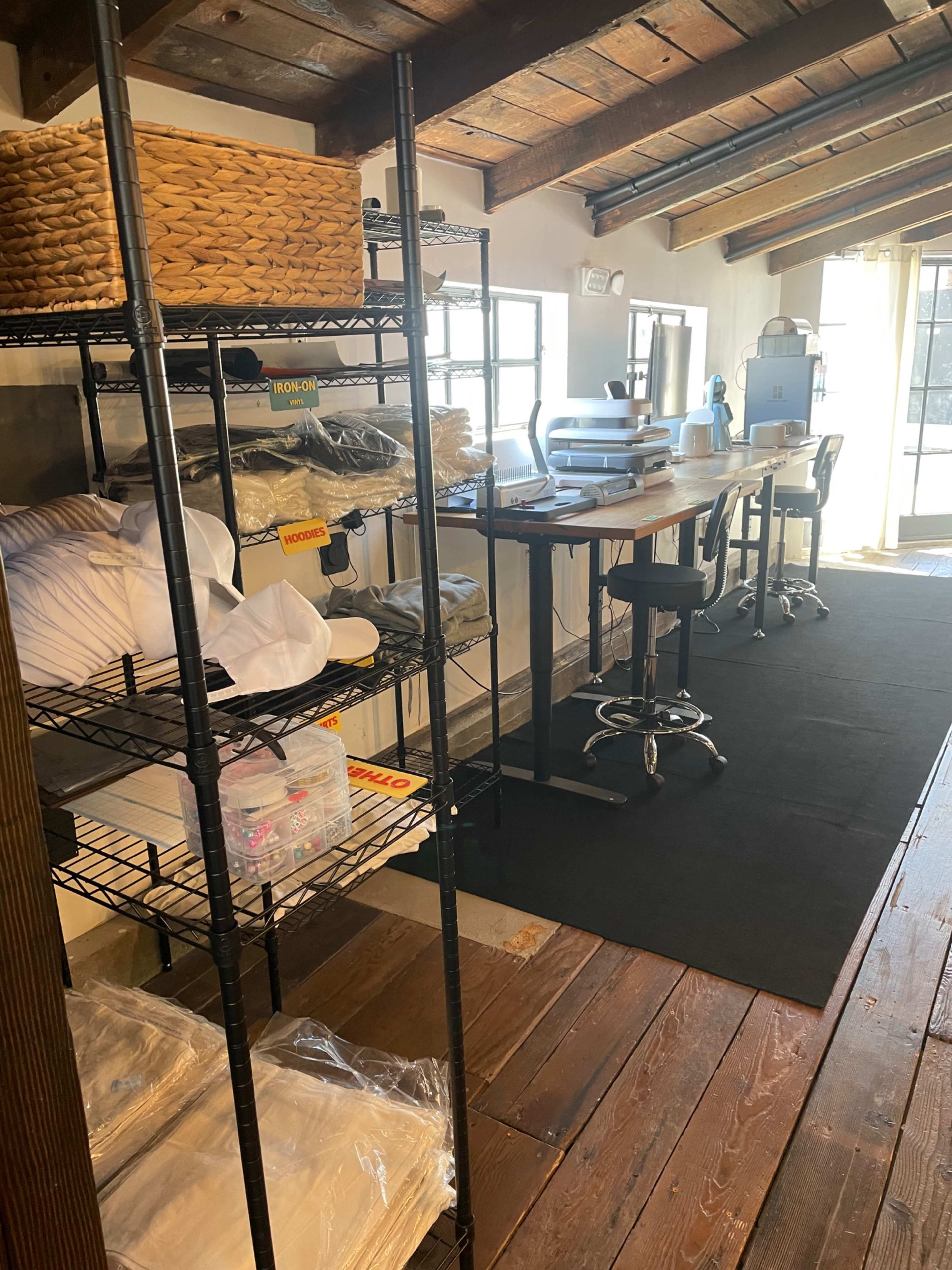 The image shows a workspace featuring shelves filled with neatly organized items, a wooden table with chairs, and a large window allowing natural light.