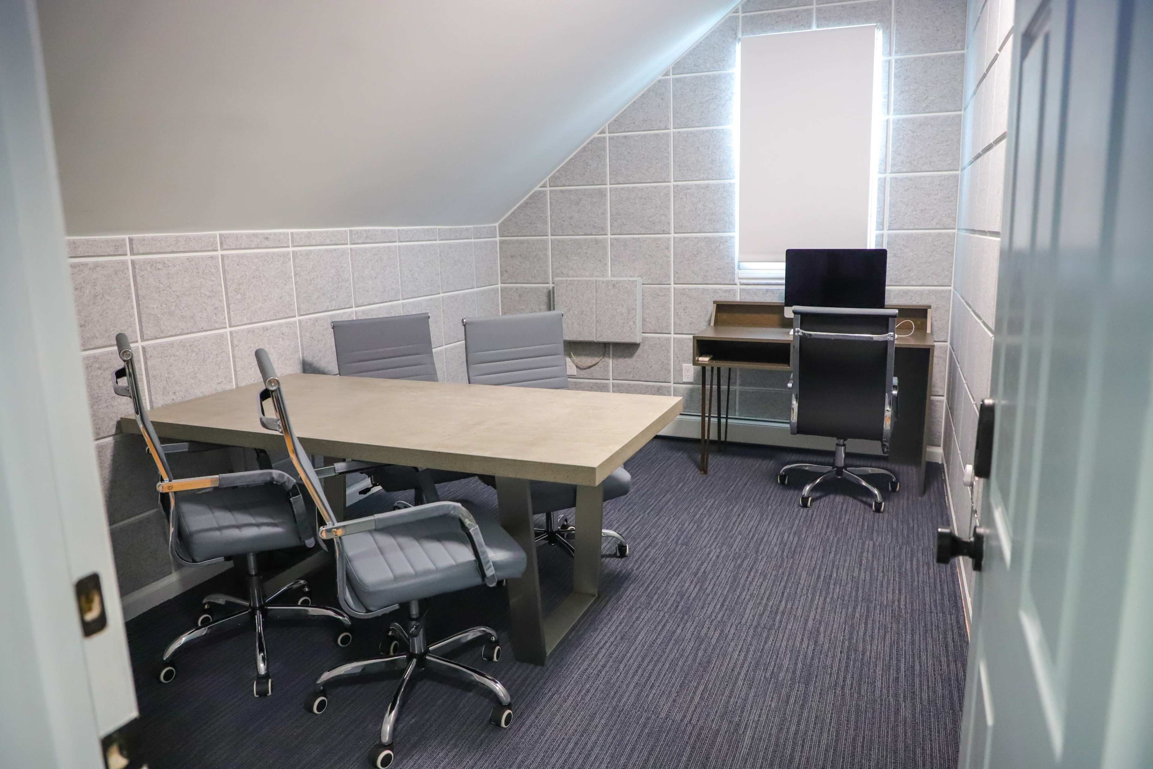 A small office space with a table, four rolling chairs, a desk, and a computer monitor against a wall with gray paneling.