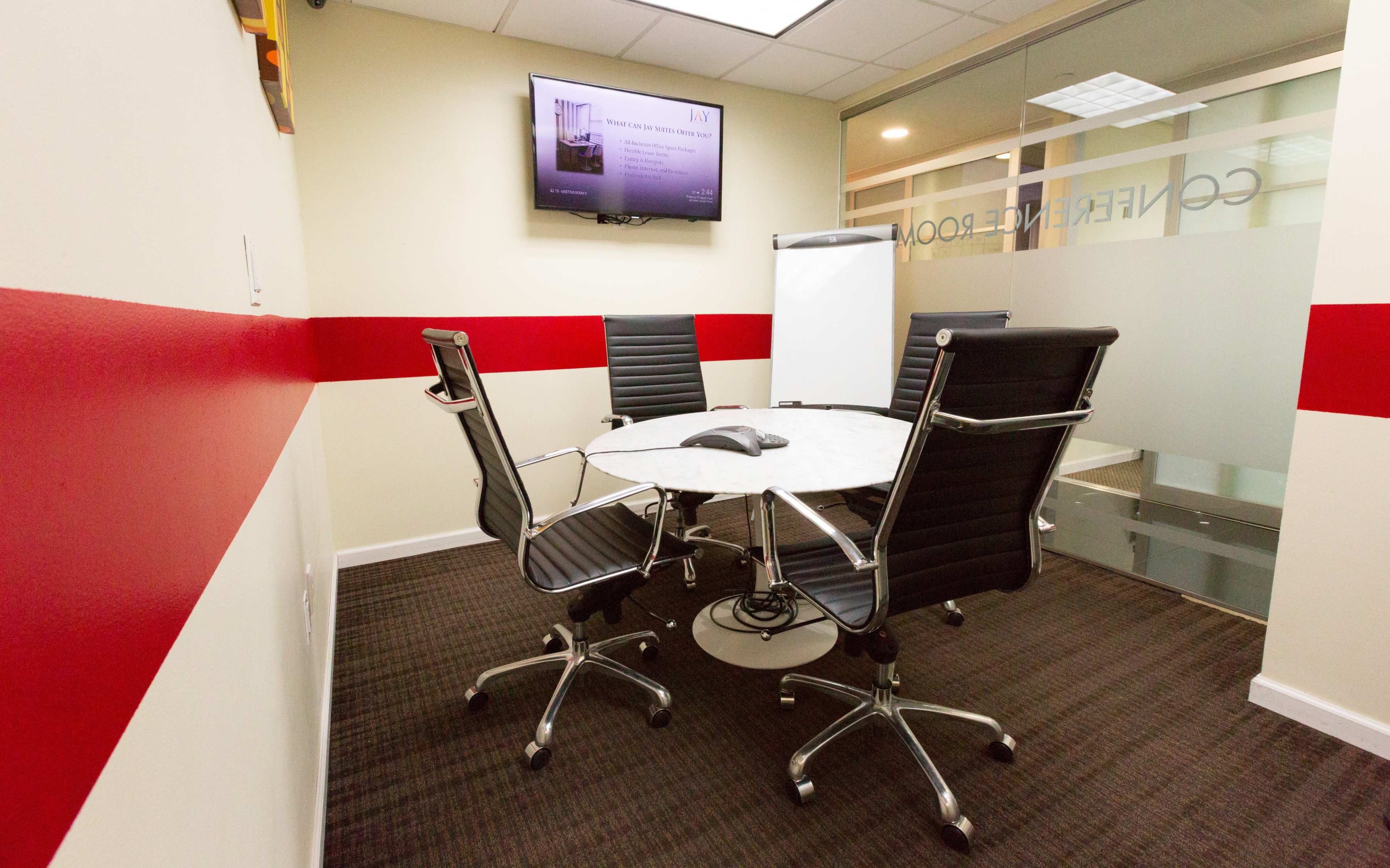 A modern conference room features a round table with four black chairs, a phone in the center, a whiteboard, and a wall-mounted TV displaying information.