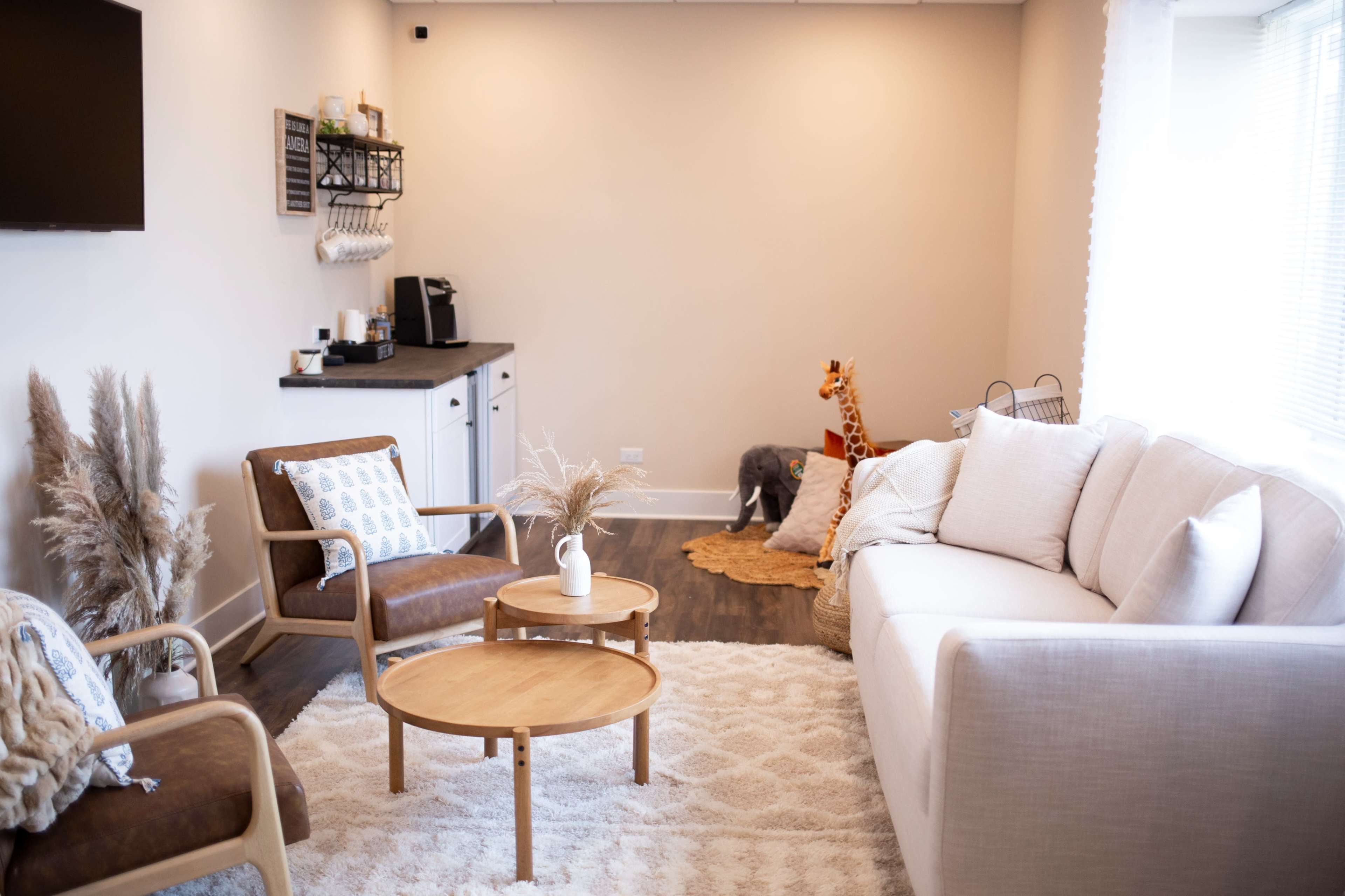 A modern living room features a light-colored sofa, wooden tables, and decorative plants, with a small kitchenette and children's toys visible in the corner.
