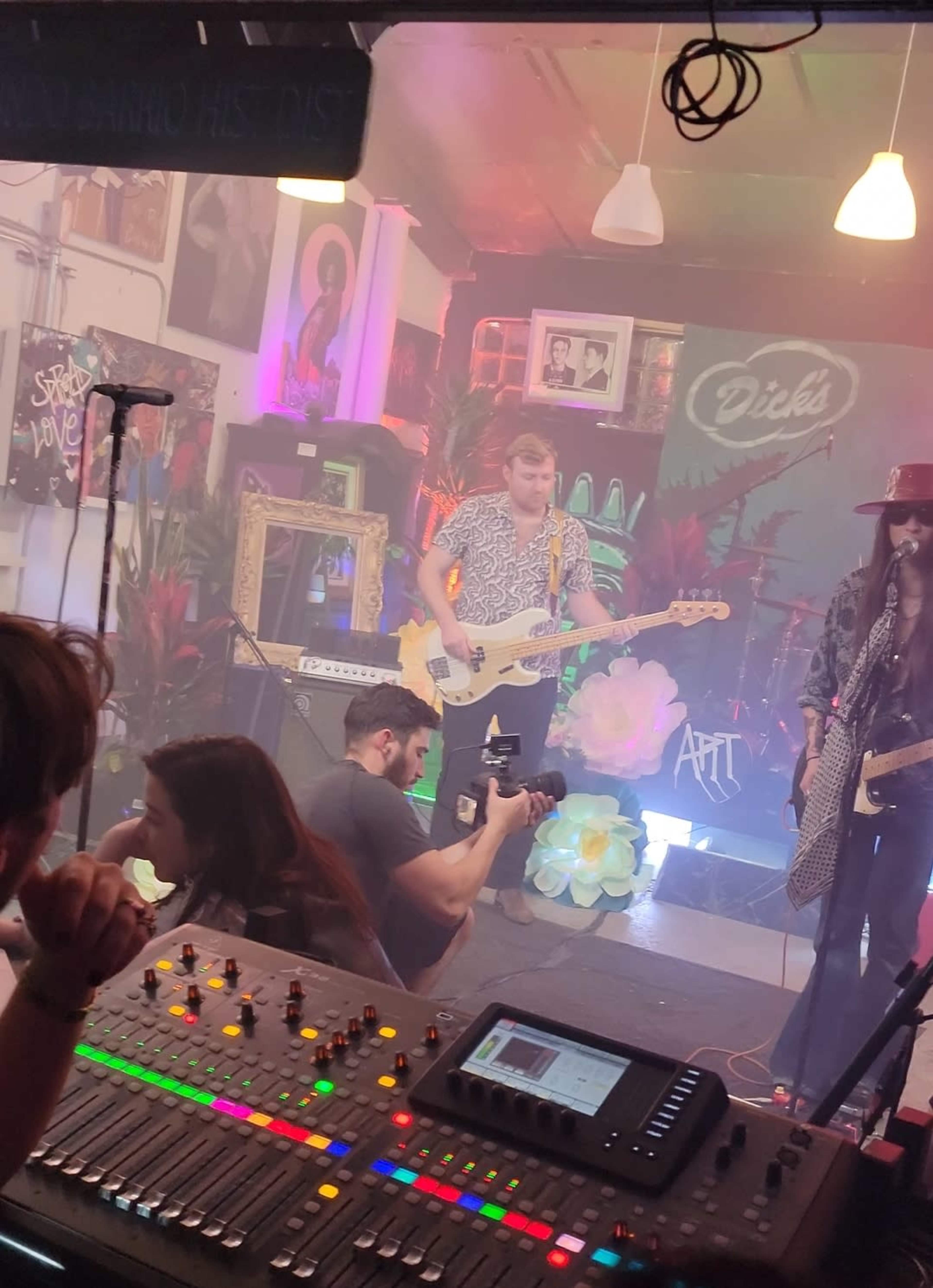 A band performs on stage while a photographer captures the moment from the audience, with a mixing board visible in the foreground.