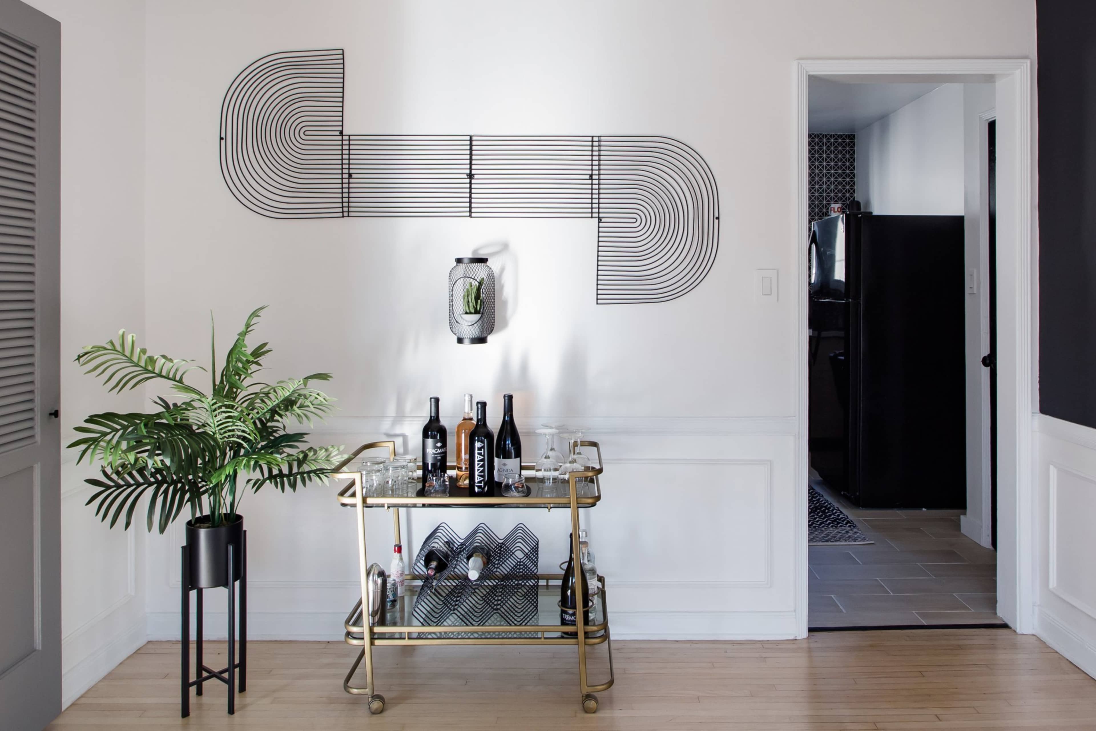 A modern bar cart with several bottles of wine and glasses is positioned in front of a wall featuring a geometric metal art piece, next to a room entrance that leads to a kitchen.