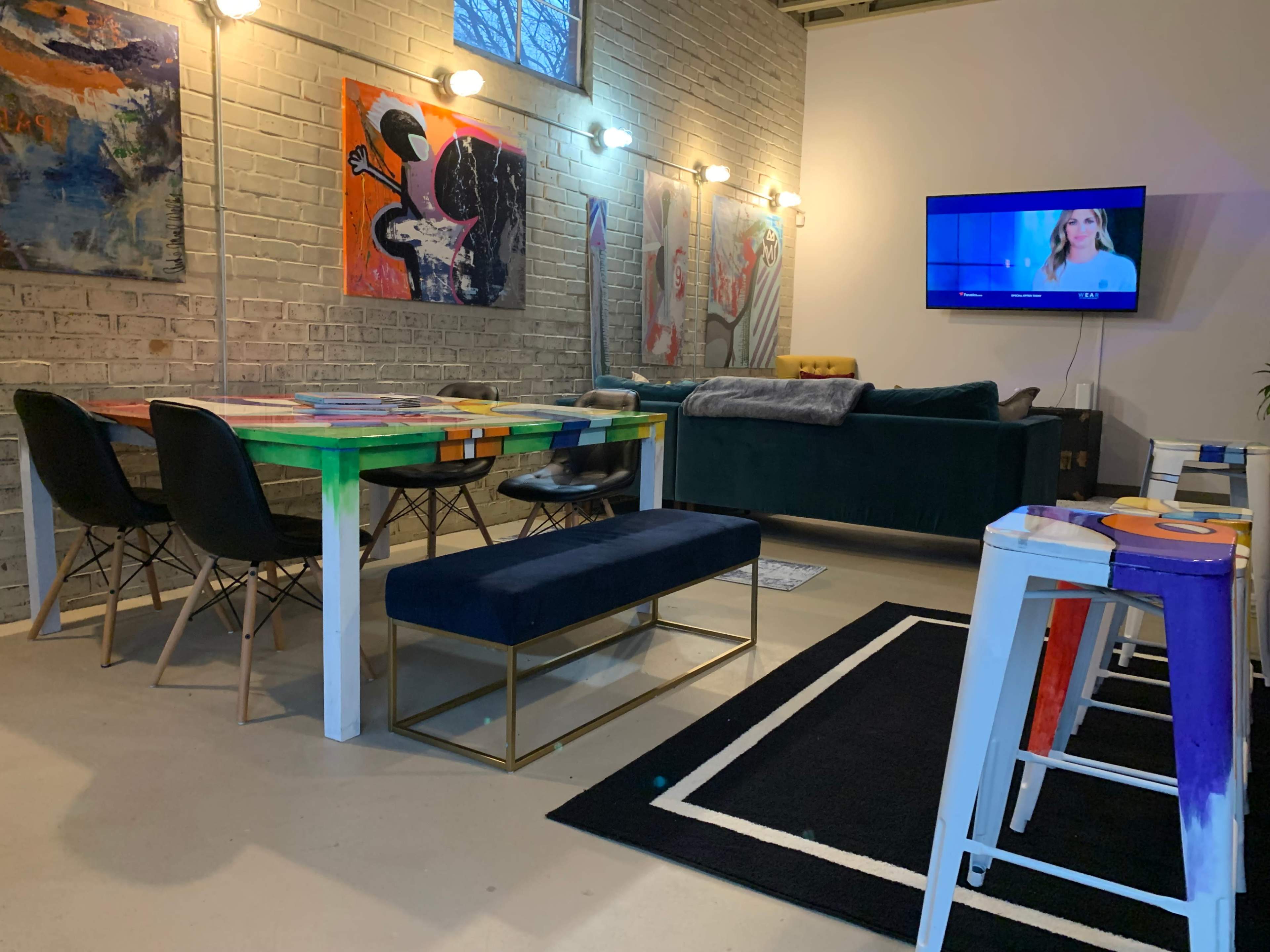 A modern living area with a dining table, chairs, a dark sofa, and a television against a white brick wall decorated with colorful artwork.