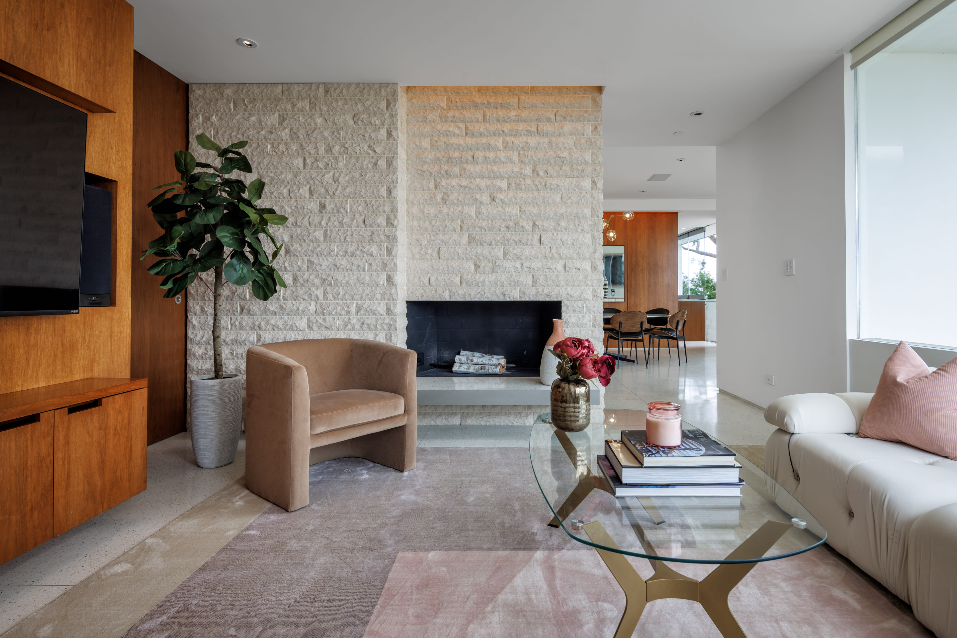 The image shows a modern living room featuring a stone fireplace, a beige armchair, a light-colored rug, a glass coffee table, and decorative items on the table.