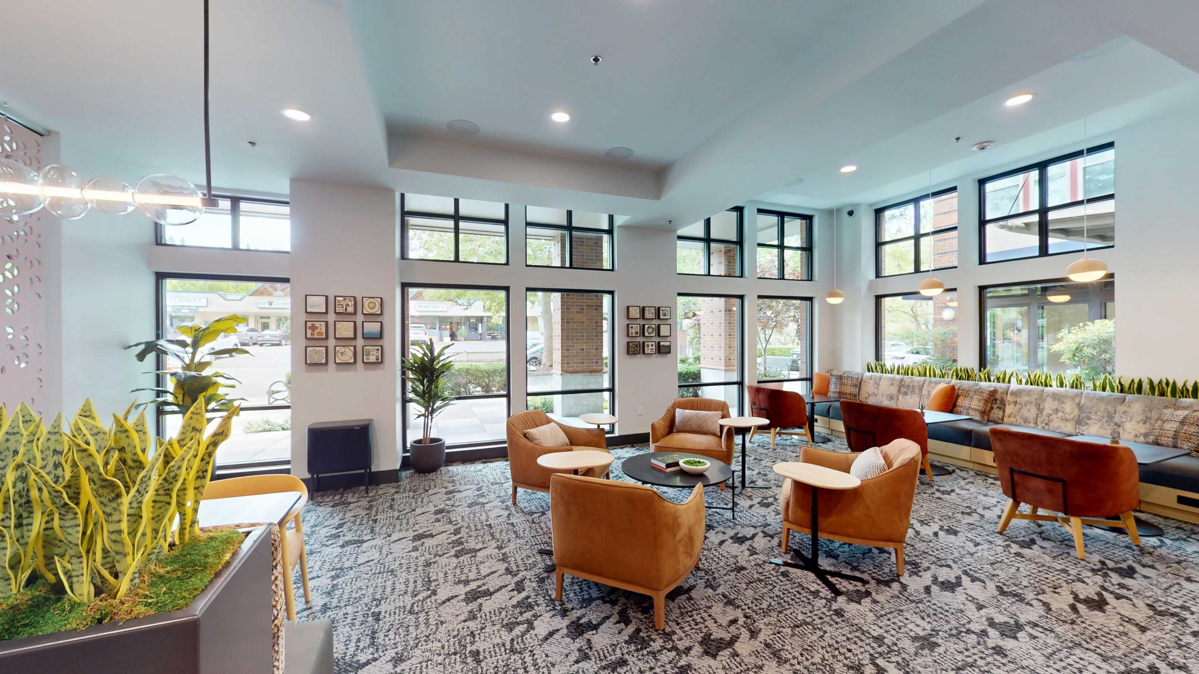 The image shows a modern lounge area furnished with comfortable chairs and a coffee table, featuring large windows that provide natural light and a view of the outdoors.
