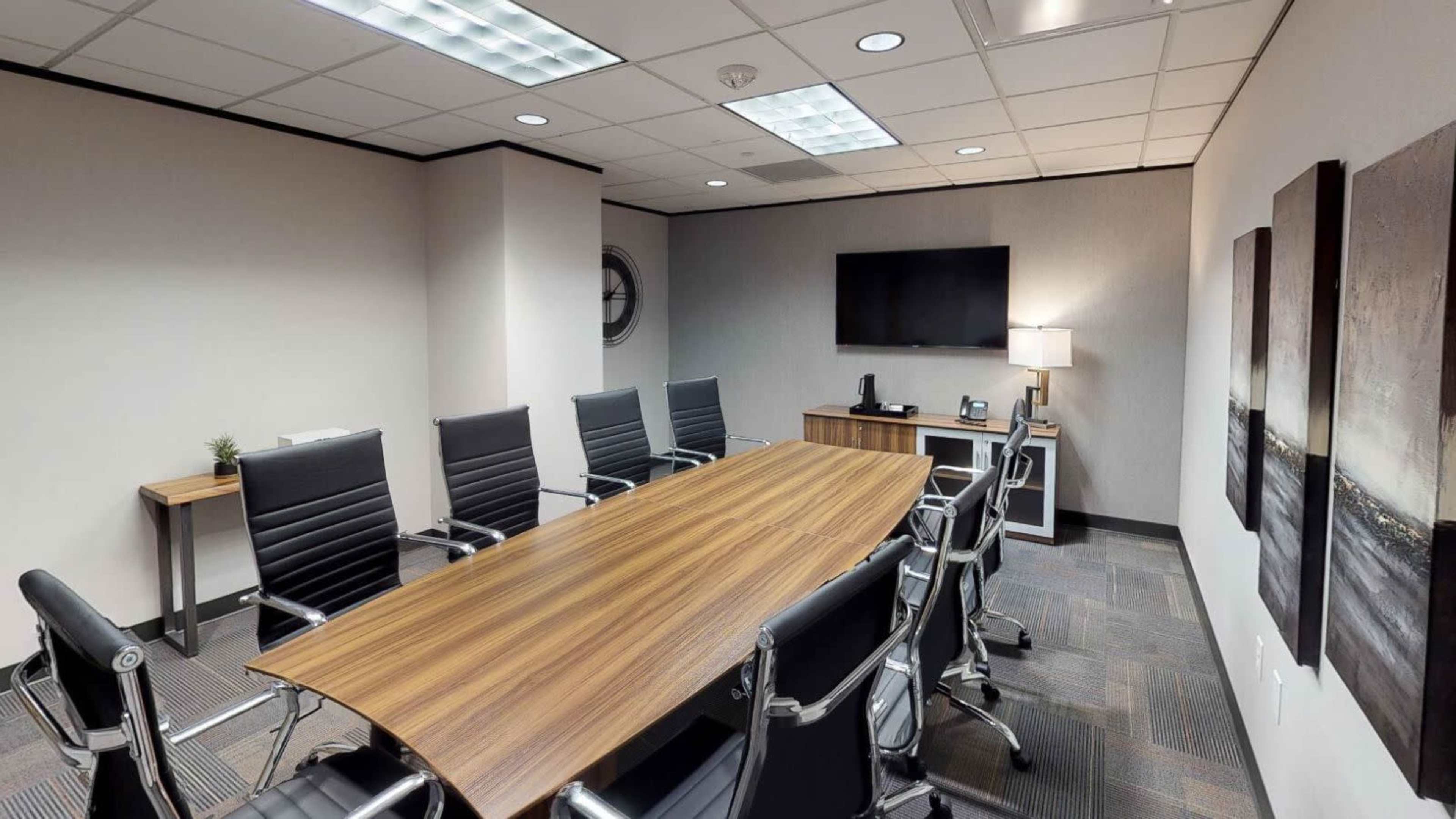 A modern conference room features a long wooden table surrounded by black leather chairs, with a television and a lamp on a side cabinet.
