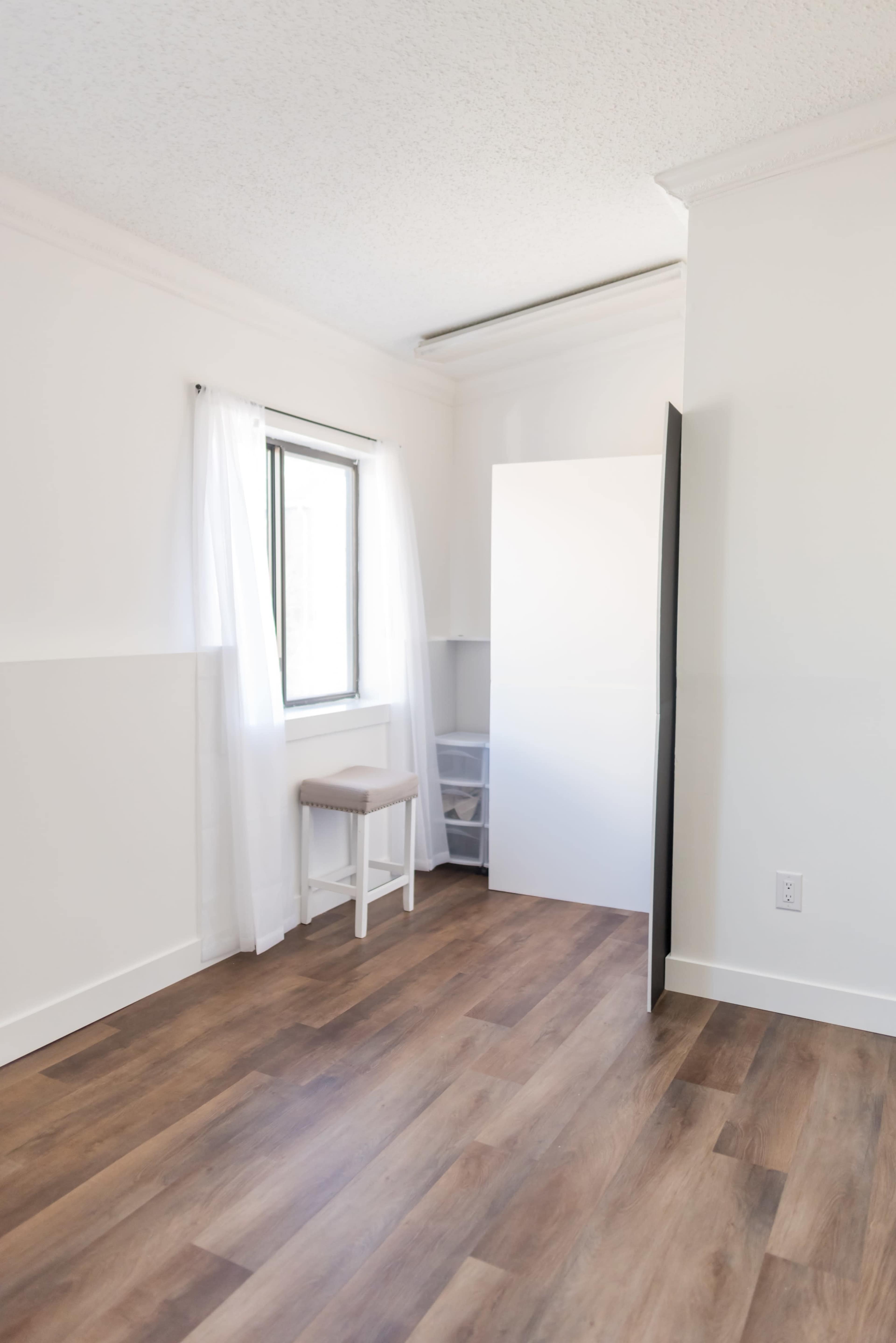 A minimalistic room with light-colored walls, a window with sheer curtains, a stool, and a white closet door.