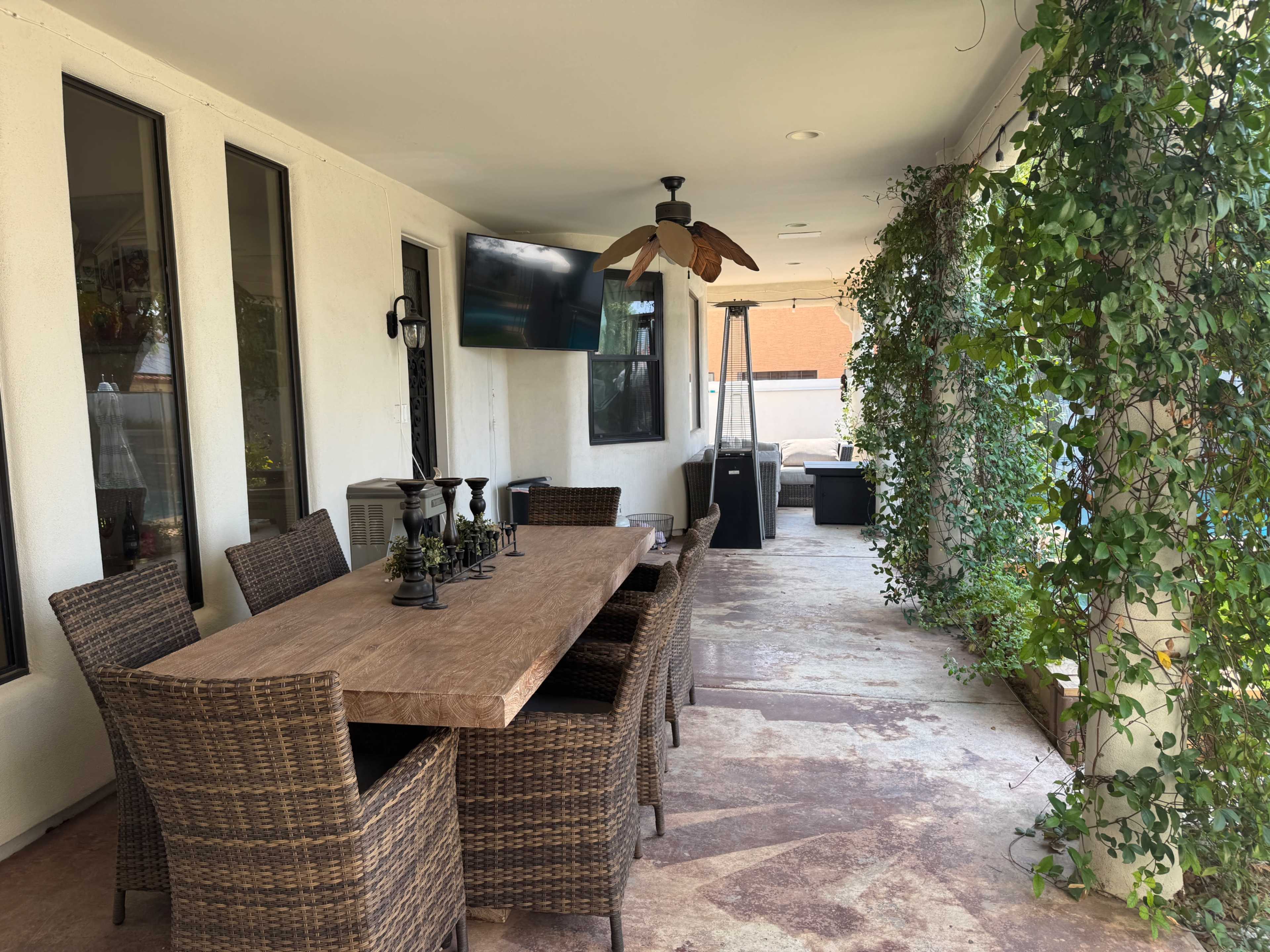 The image shows a covered outdoor patio featuring a large wooden dining table surrounded by wicker chairs, with a television mounted on the wall and greenery along one side.