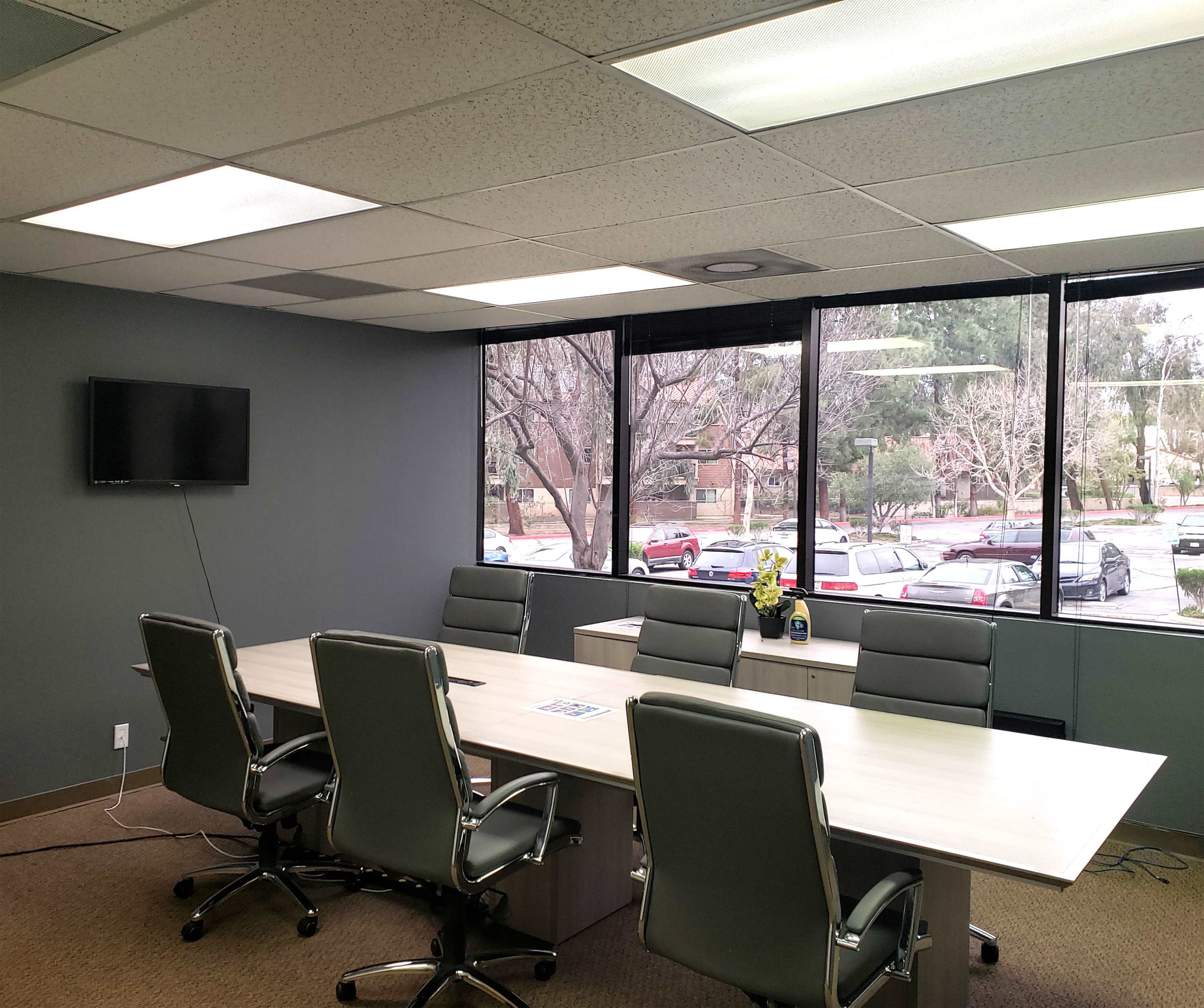 A modern conference room features a long table surrounded by ergonomic chairs, with large windows overlooking a parking lot and trees.