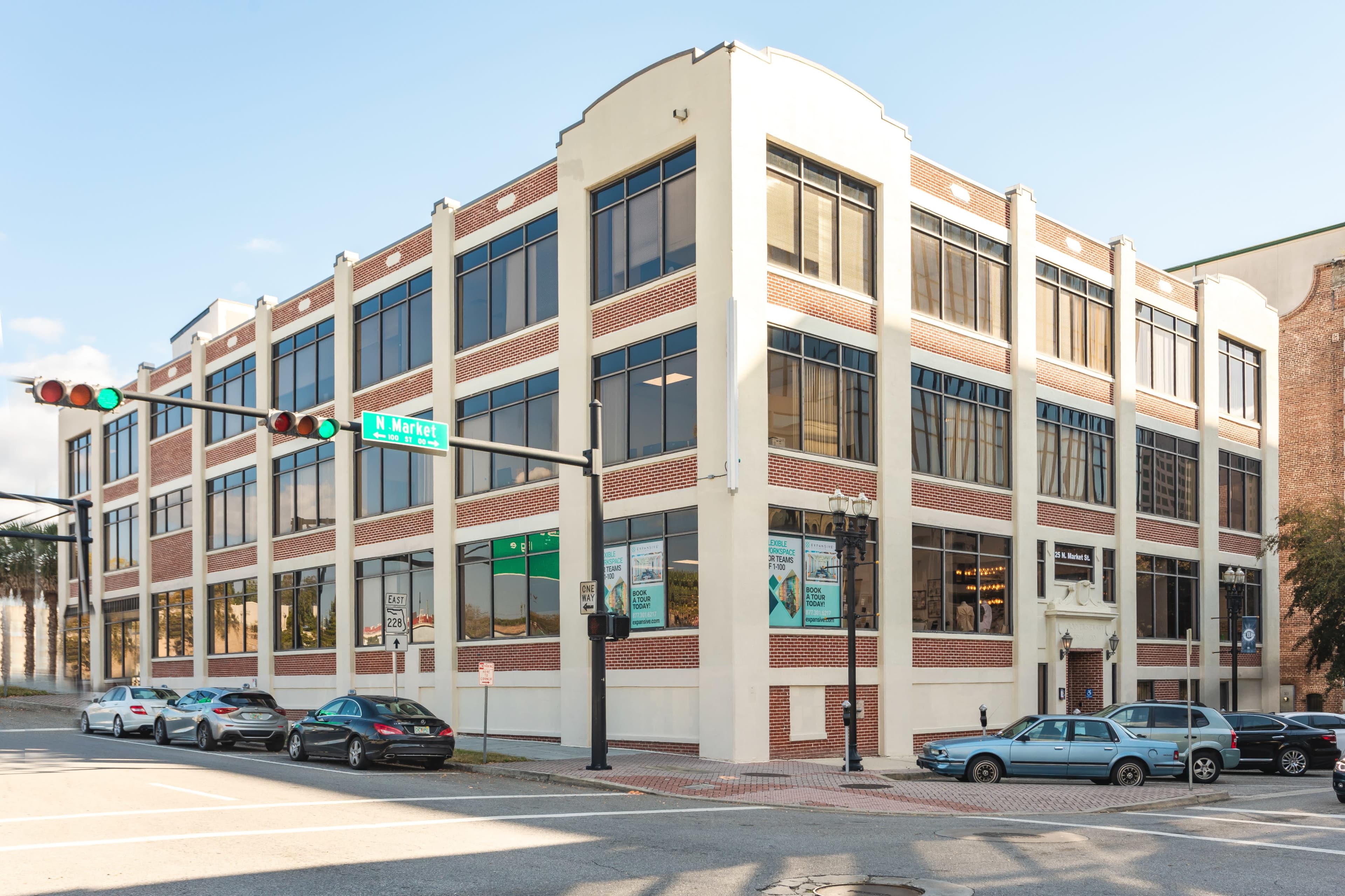 The image shows a multi-story building with a mix of glass and brick facade, located at the corner of a street with parked cars in front.