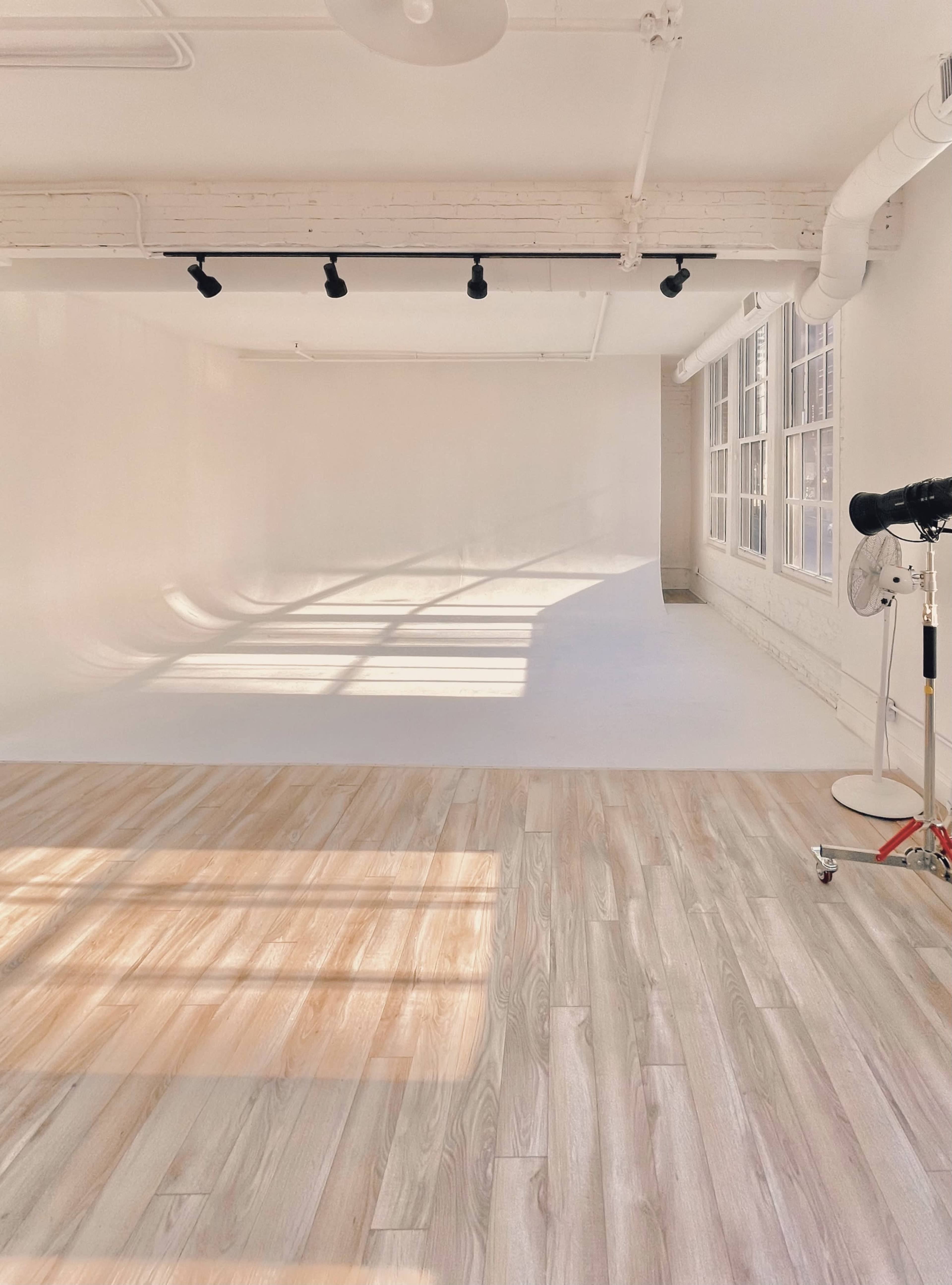 A spacious white studio with a curved backdrop and wooden flooring, illuminated by sunlight filtering through large windows.