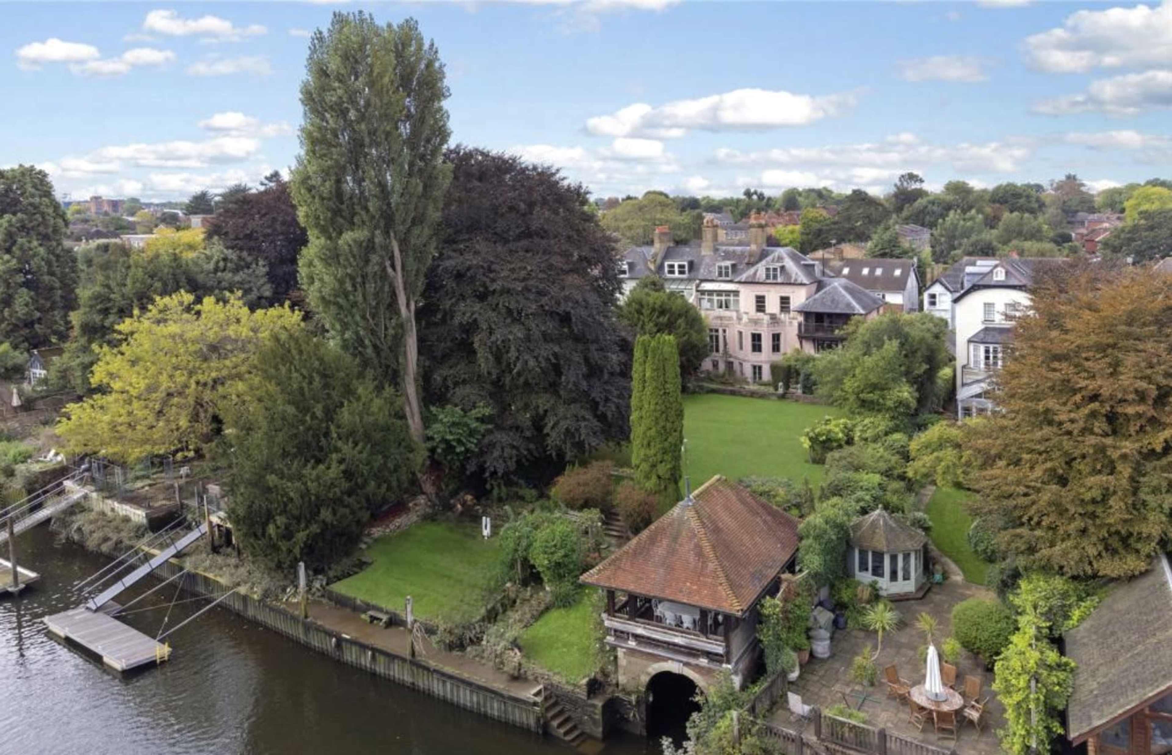 The image shows a landscaped garden with multiple trees and a gazebo beside a river, along with several houses in the background.