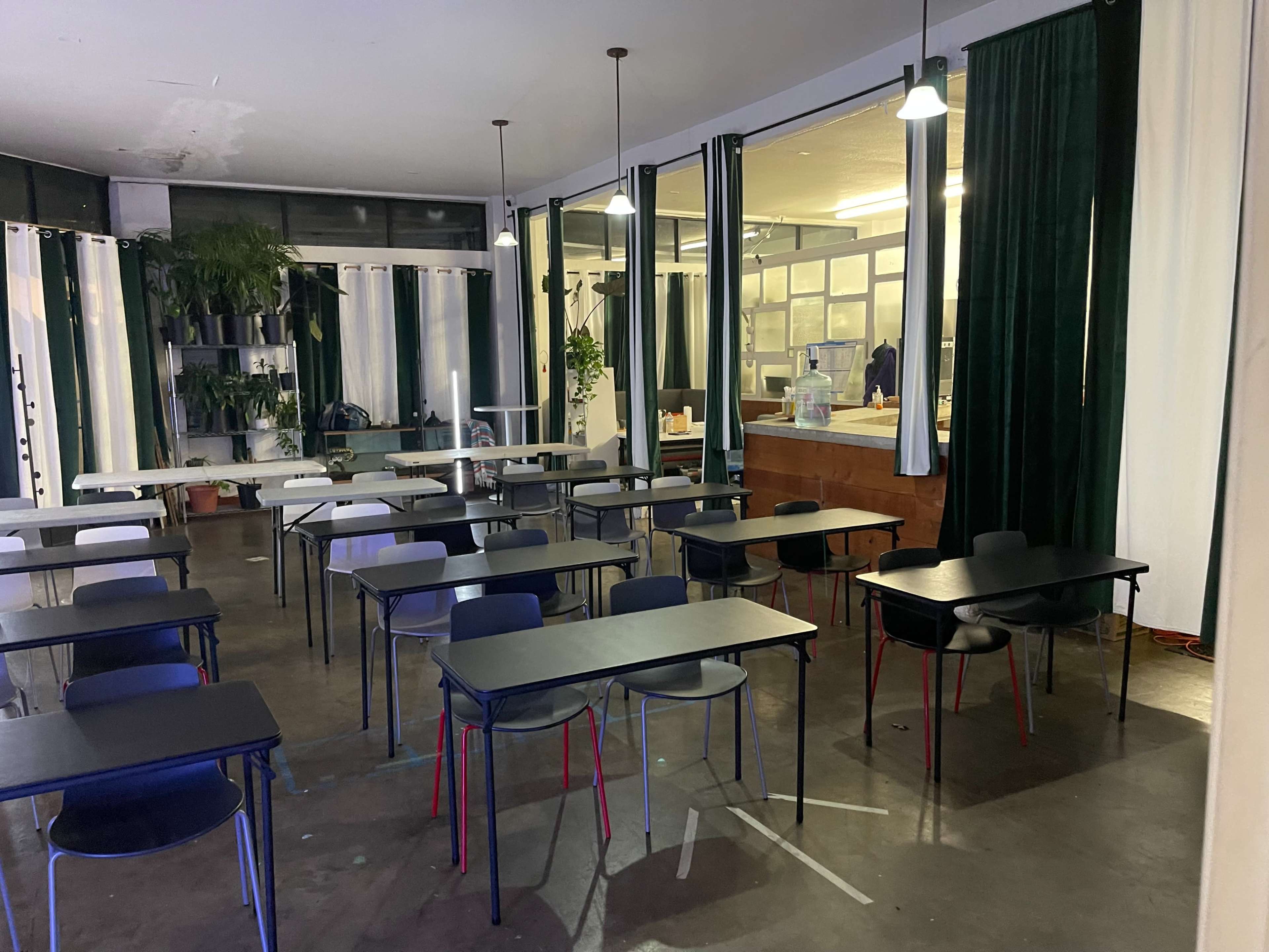 A classroom is set up with several tables and chairs, featuring green curtains and potted plants in the background.