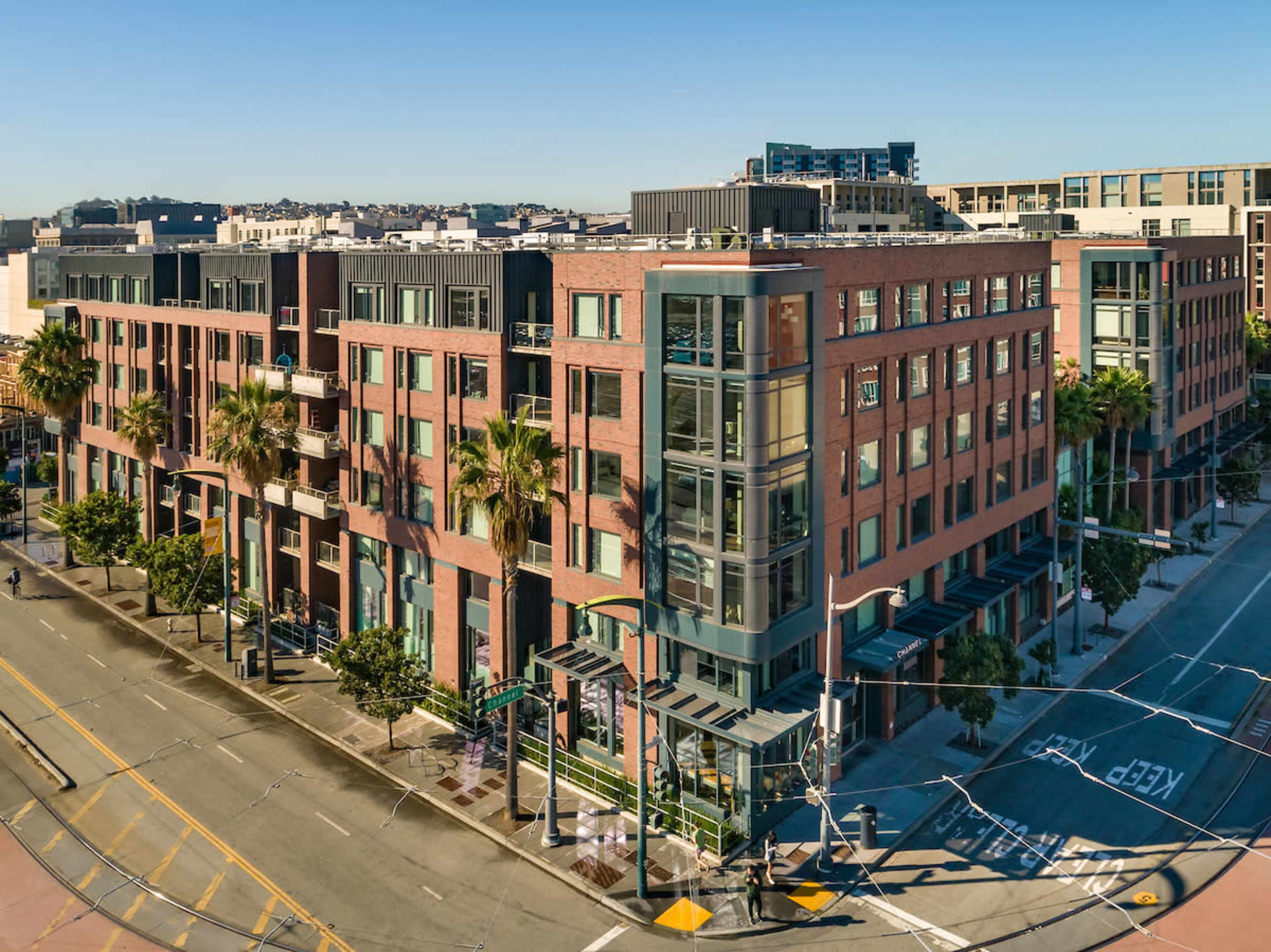 A modern brick apartment building with large windows and palm trees lining the street at a busy intersection.