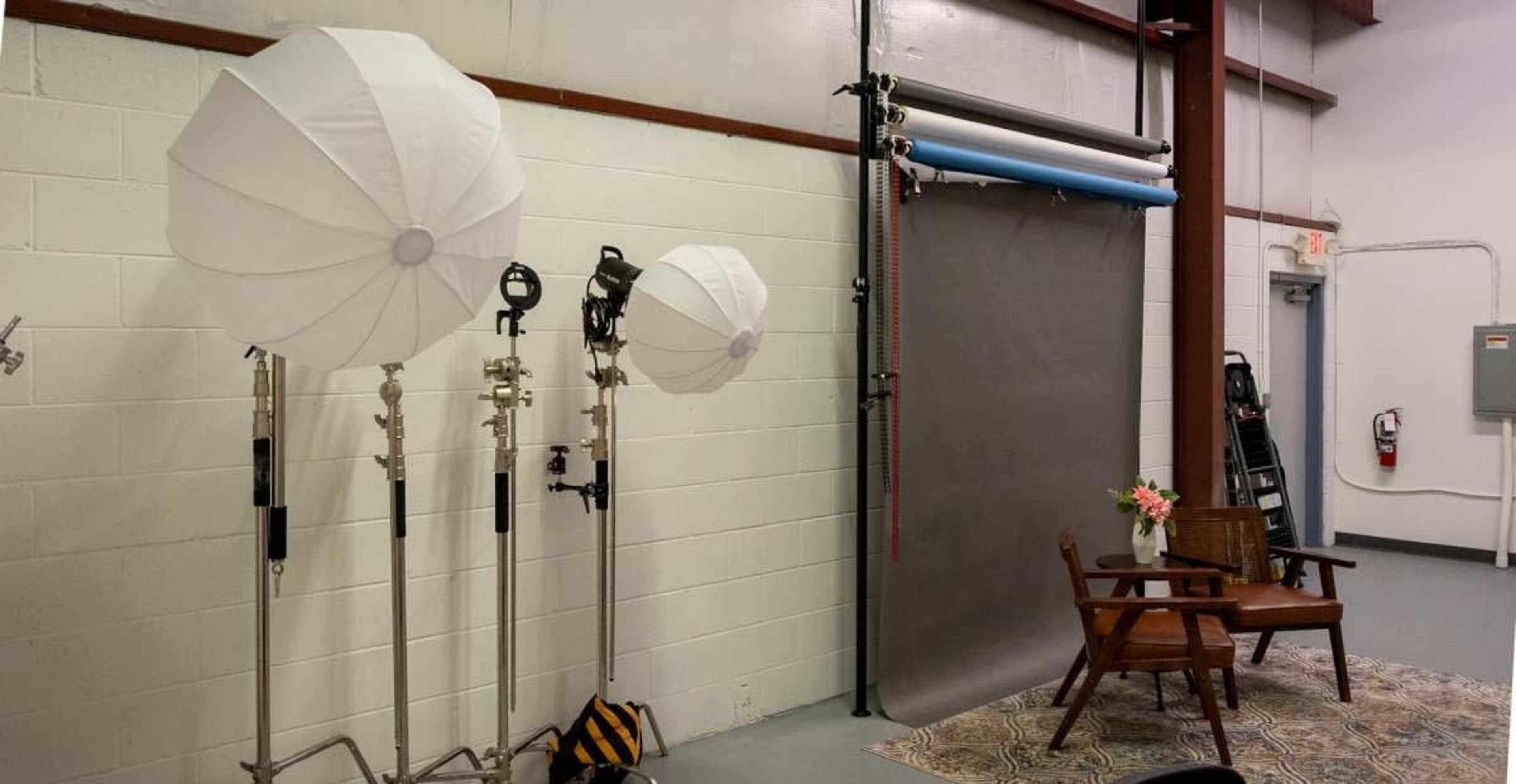 The image shows a photography studio with three large softbox lights, a gray backdrops, and a wooden chair with a small vase of flowers on a patterned rug.