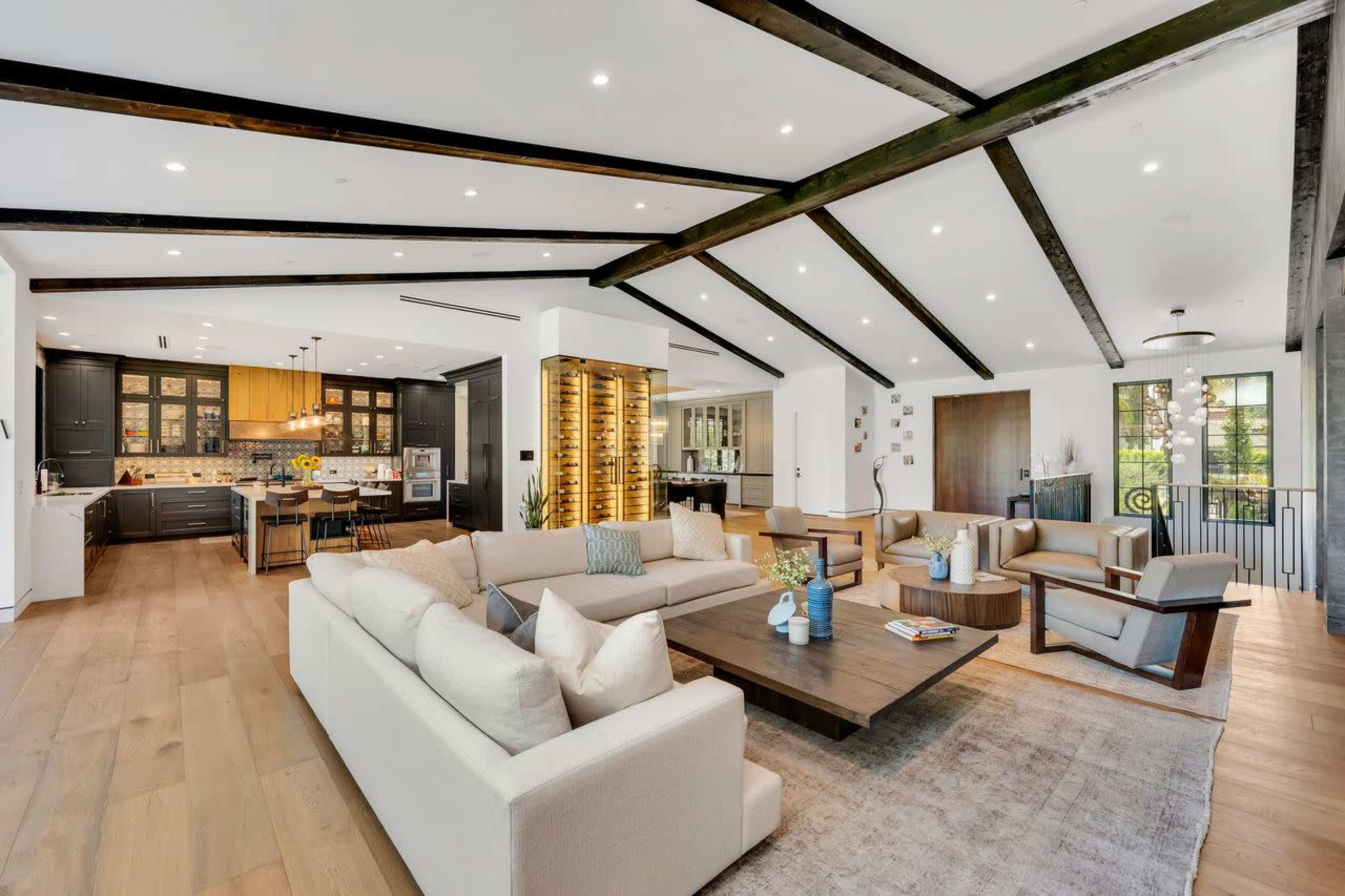The image shows a modern living room with a large sectional sofa, a wooden coffee table, and exposed beam ceilings, opening up to a kitchen area with dark cabinetry.