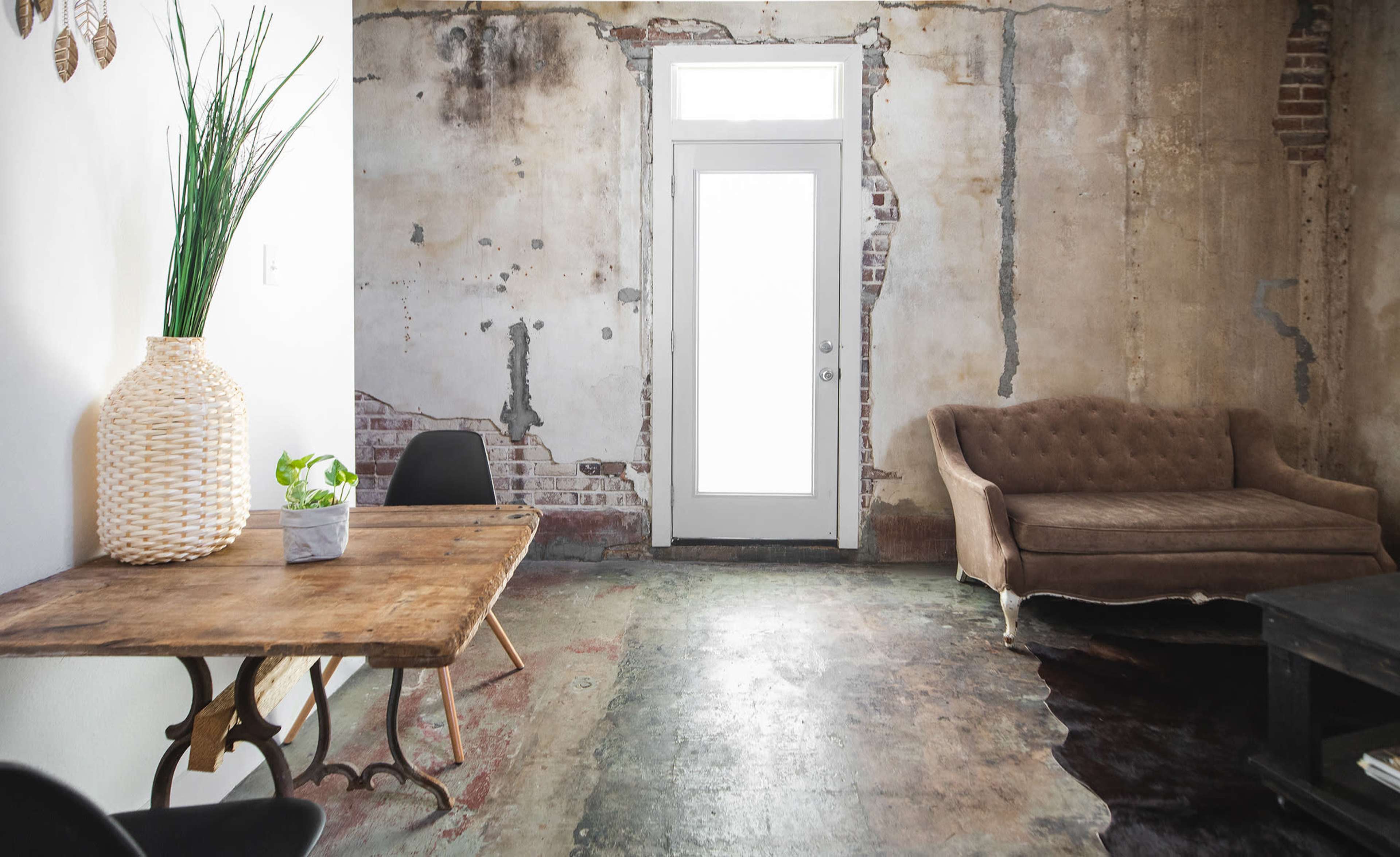 A room with a weathered wall, a wooden table, a black chair, a beige sofa, and a door leading outside.