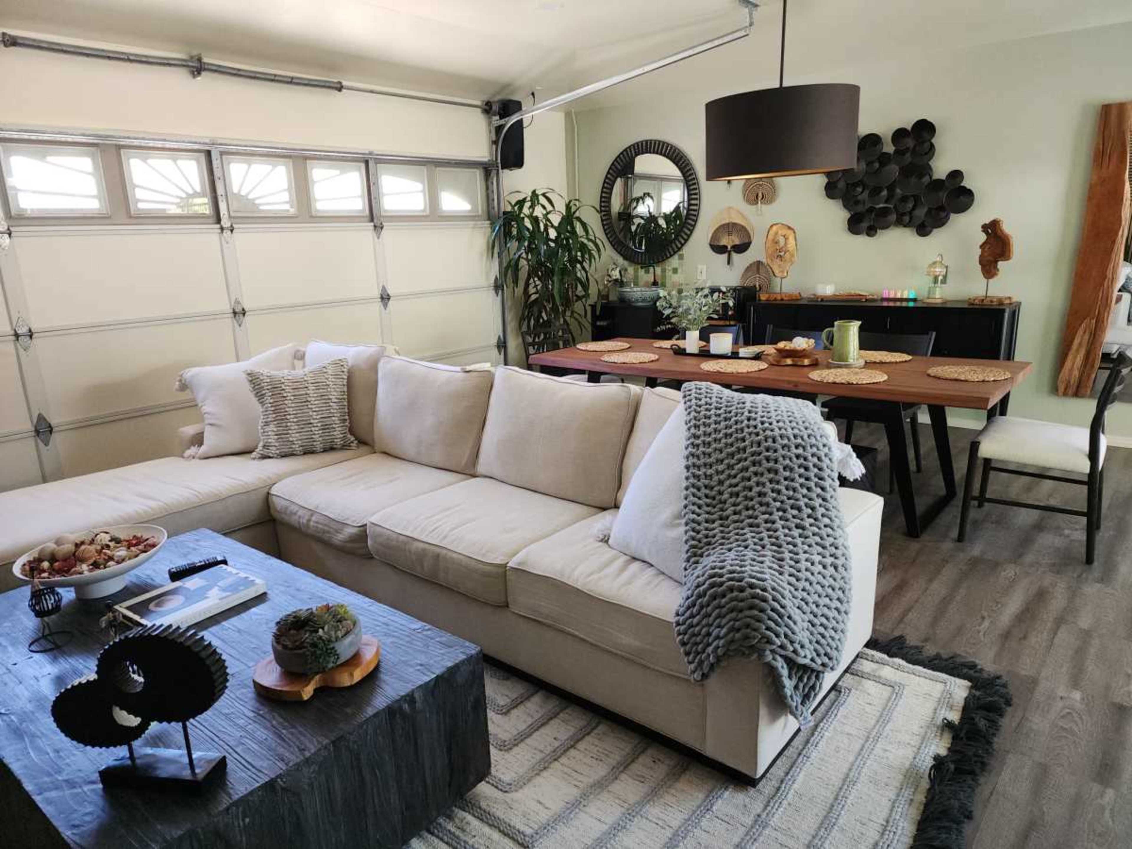 The image shows a modern living space featuring a beige sectional sofa, a dark wooden coffee table, and a dining area with a large table and decorative items on the walls.