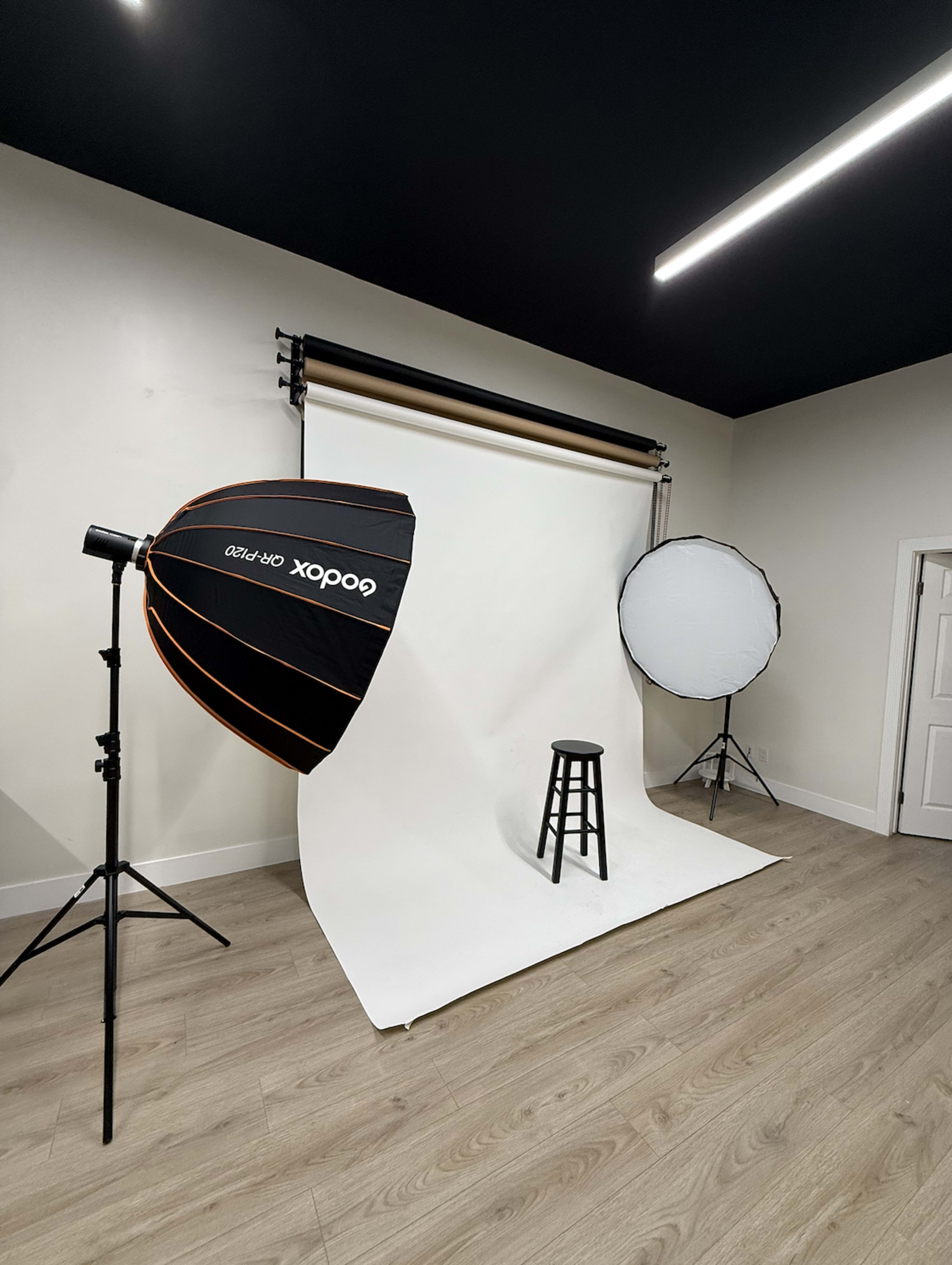 The image shows a photography studio setup featuring a light umbrella, a reflector, and a wooden stool positioned in front of a white backdrop.