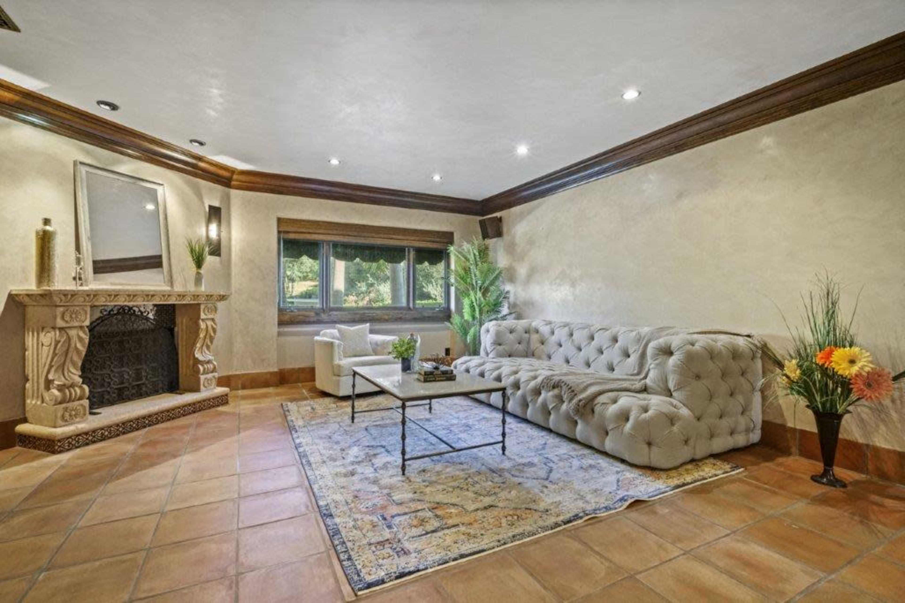 The image shows a living room with a large, tufted sectional couch, a coffee table, a single white armchair, and a stone fireplace, all against a backdrop of warm-toned walls and flooring.