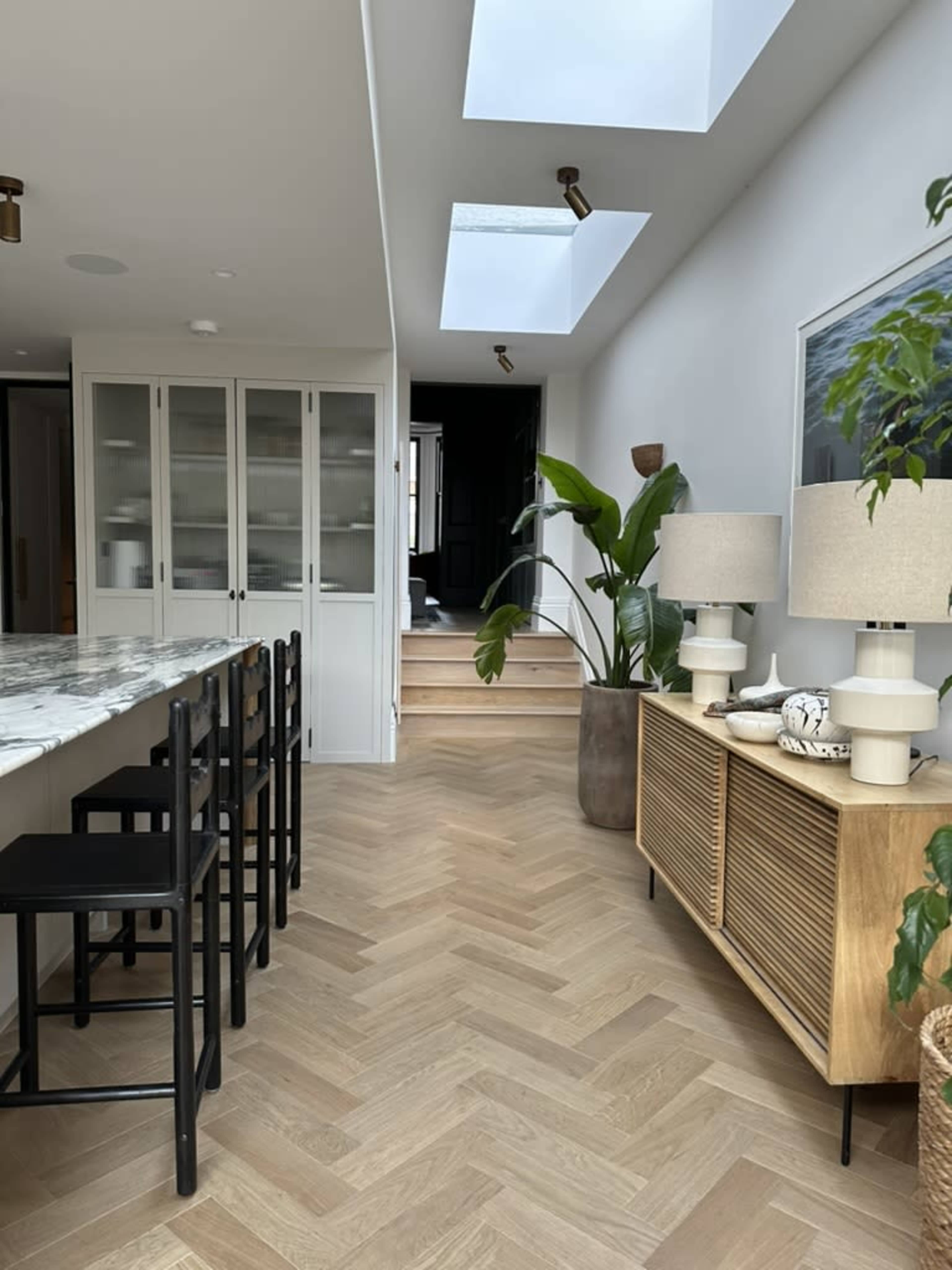 The image shows a modern interior with a herringbone wood floor, a kitchen island with black bar stools, and a wooden sideboard next to a large green plant, leading to a stairway and open skylights above.