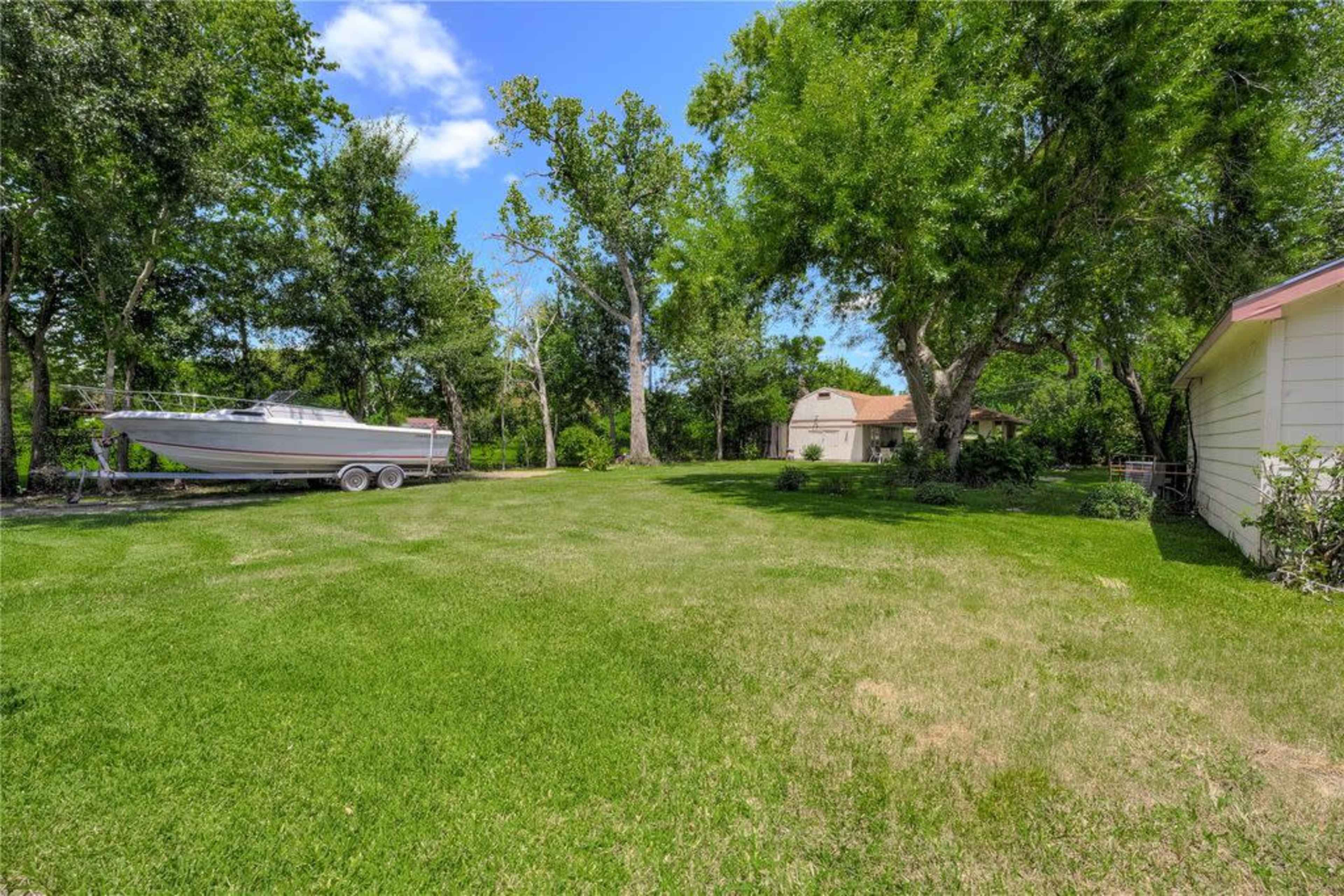 The image shows a grassy backyard with a parked boat on a trailer and a shed in the background surrounded by trees.