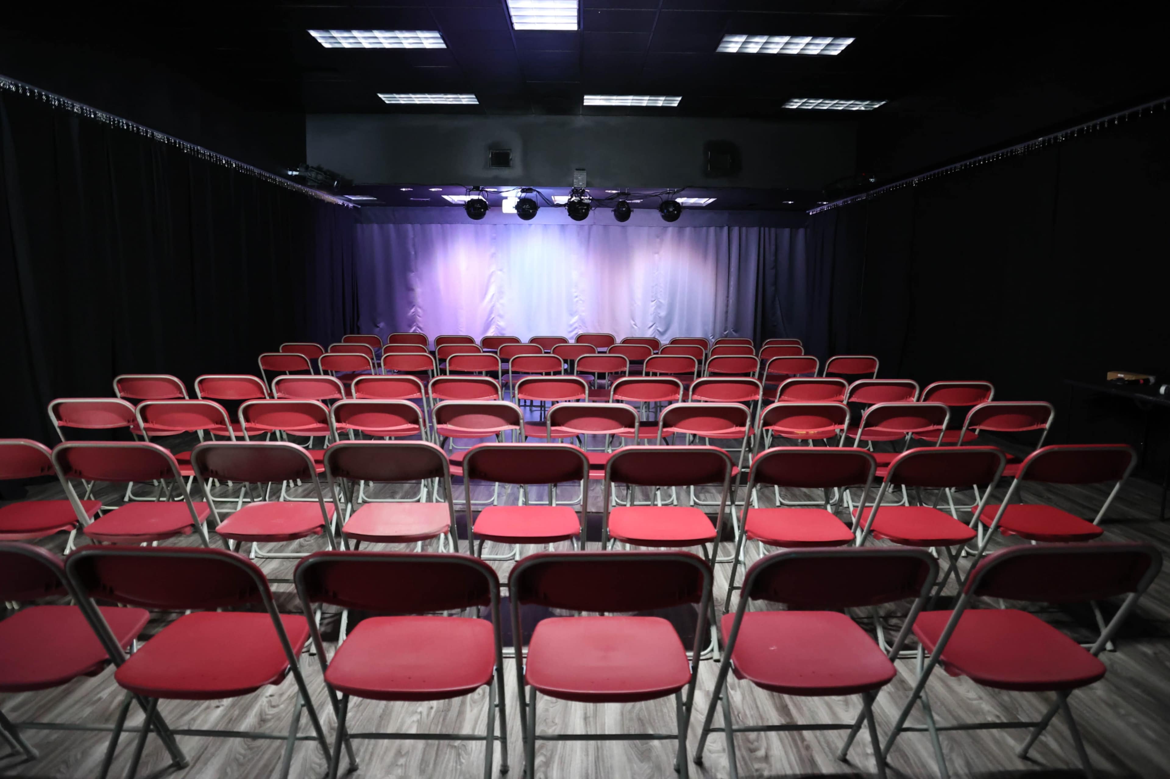 A theater with rows of red chairs facing a stage illuminated by overhead lights.