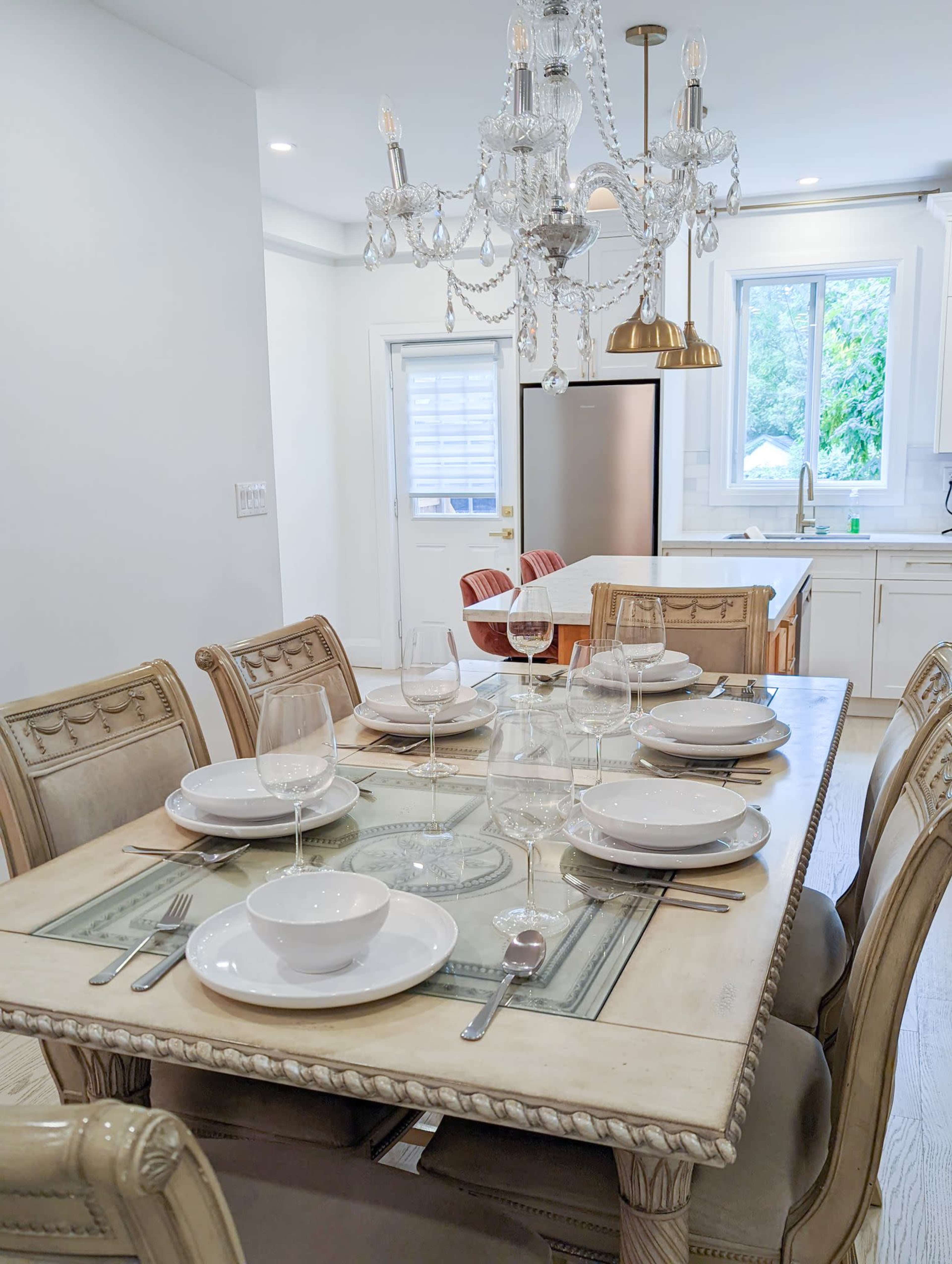 A dining table is set with plates, bowls, and glasses in a well-lit kitchen featuring a chandelier and modern appliances.
