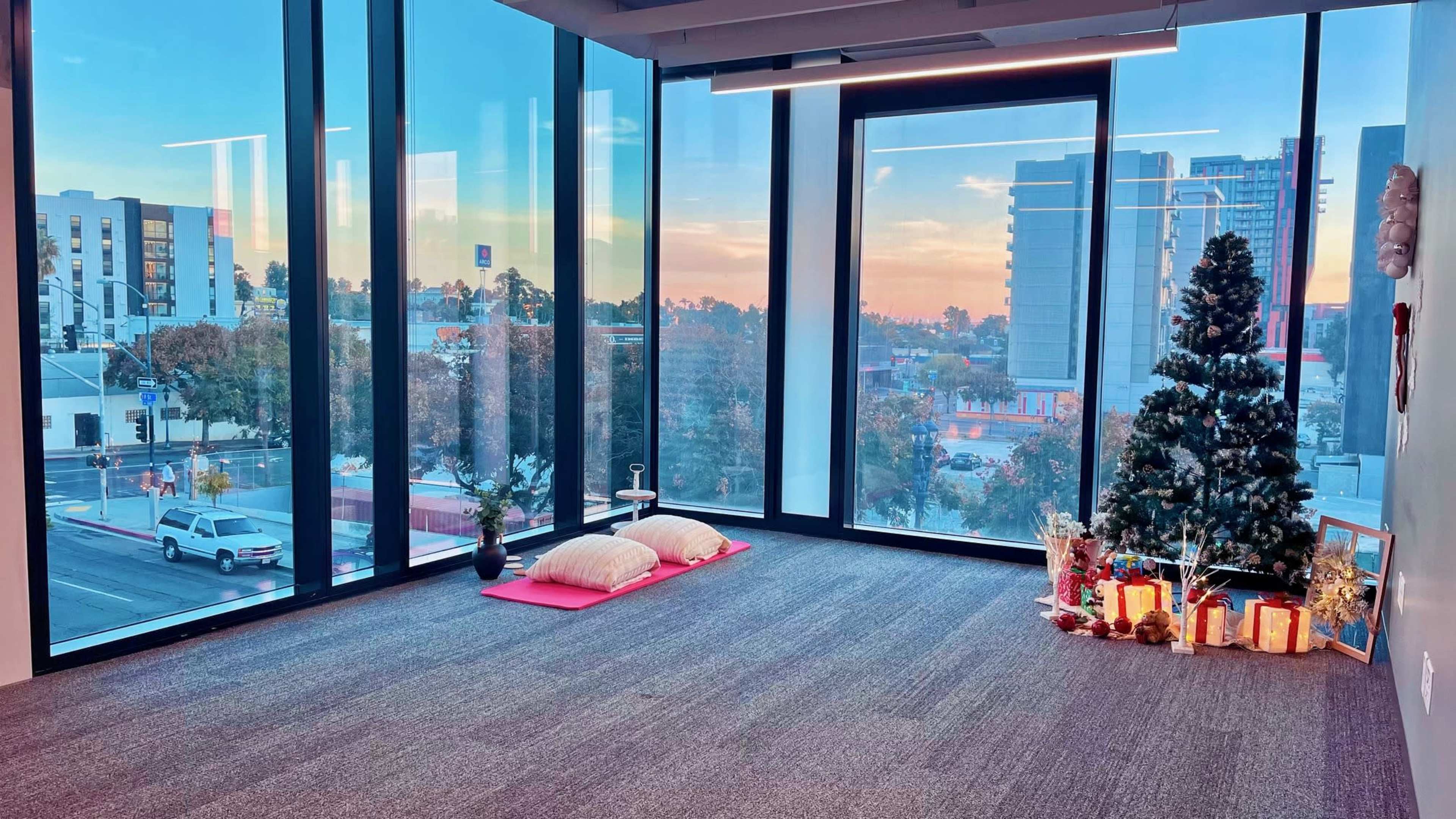An empty room with large windows displaying a city skyline at sunset, featuring two floor cushions and a decorated Christmas tree in one corner.