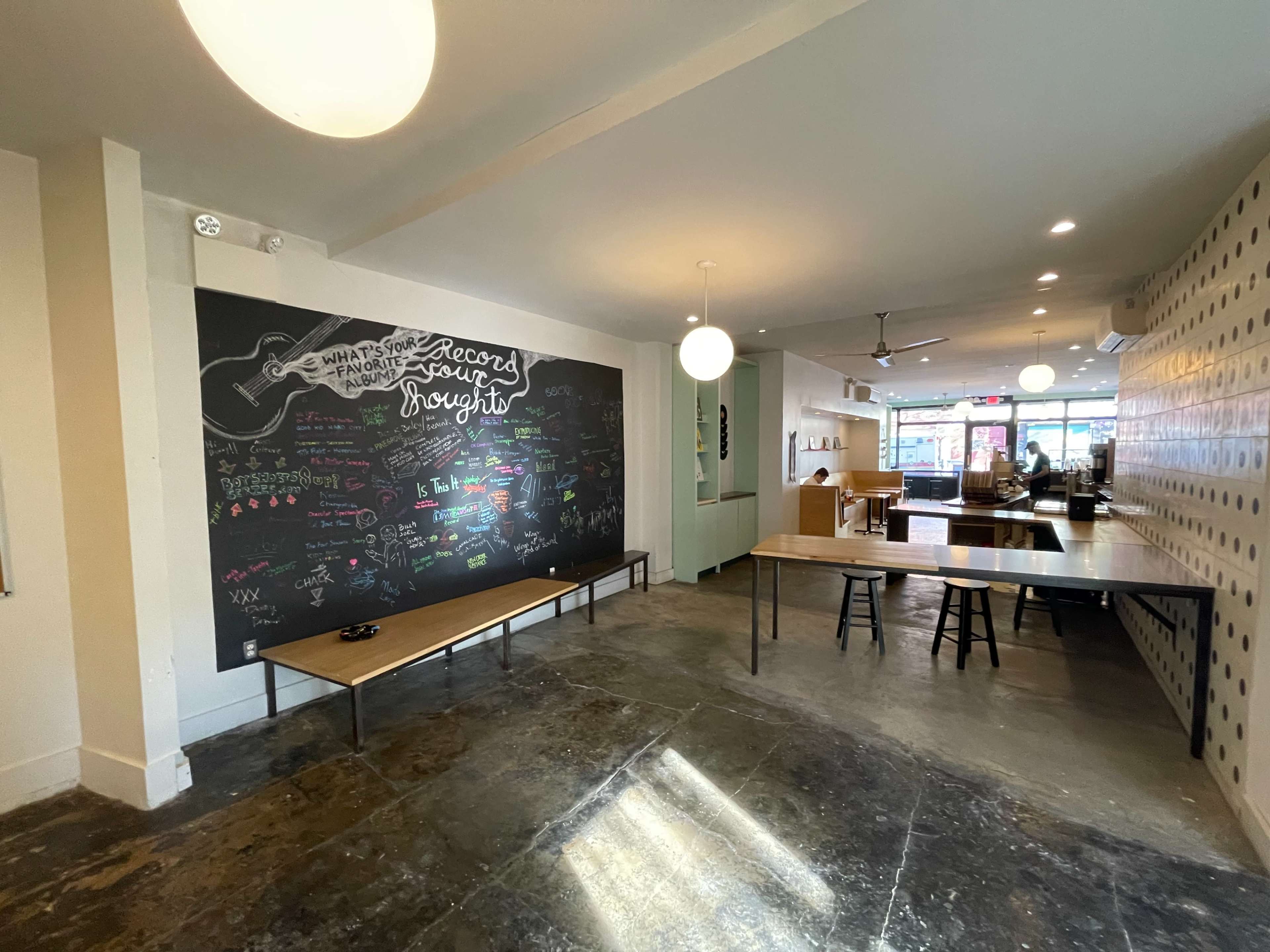 A spacious café interior featuring a large chalkboard wall with colorful writing and two wooden tables set up near the floor.