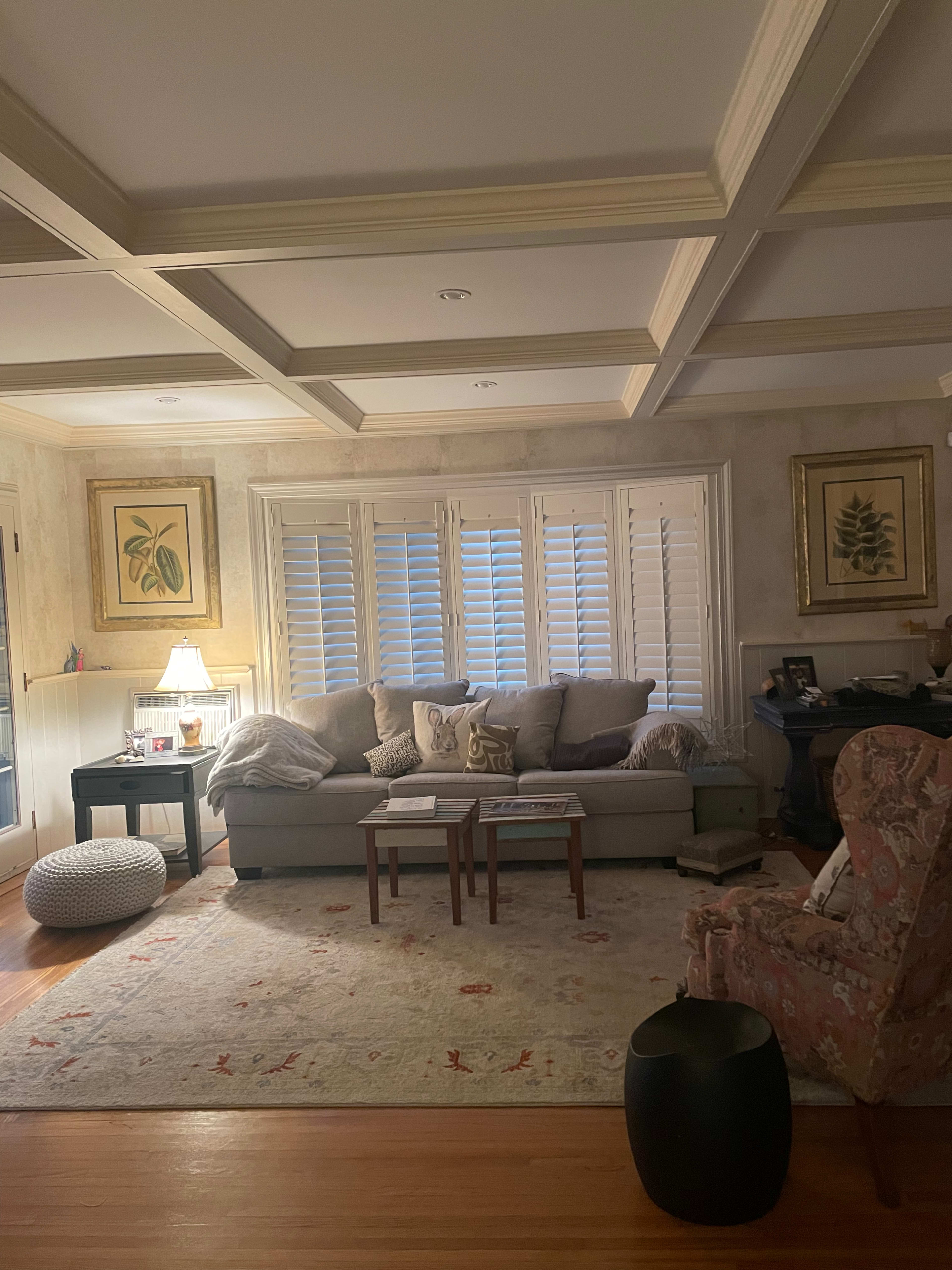A cozy living room features a light gray sectional sofa, a wooden coffee table, and a patterned area rug, with light filtering through plantation shutters.