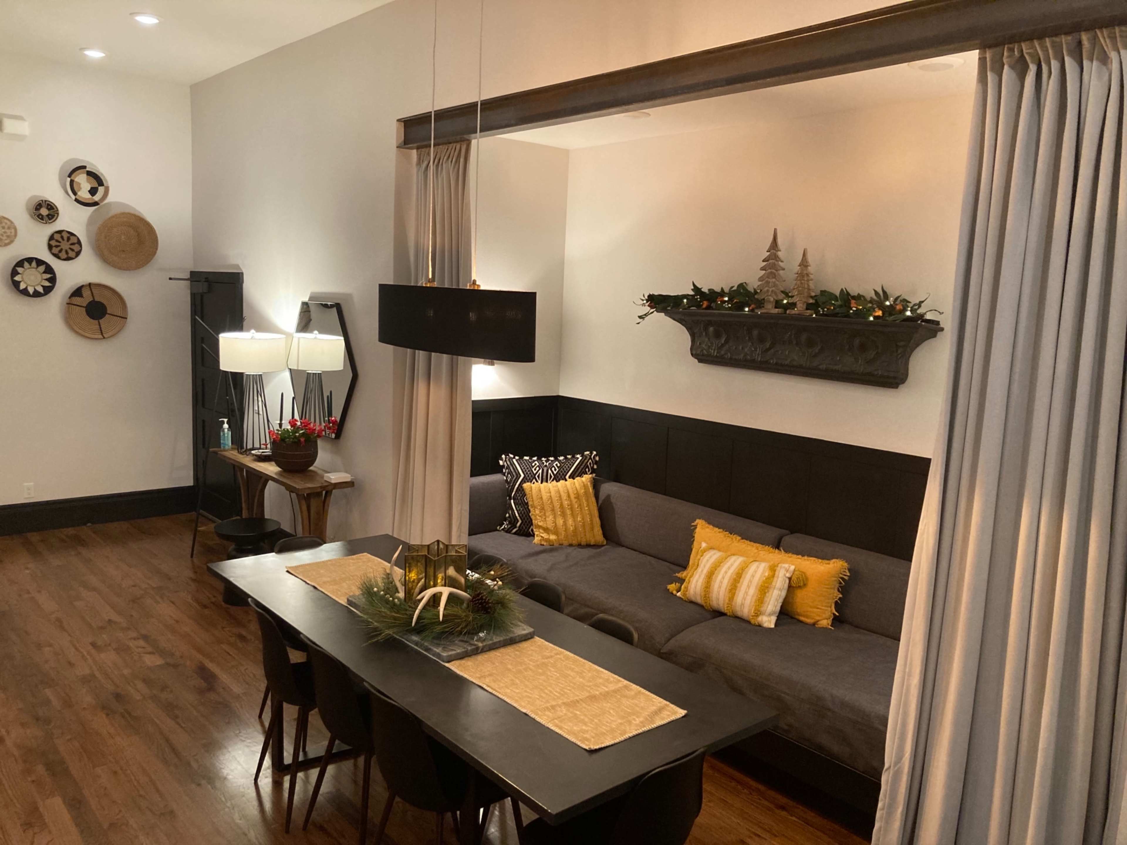 The image shows a living space featuring a gray sofa with yellow accent pillows, a dining table set for meals, and decorative elements including a holiday-themed centerpiece and wall decor.