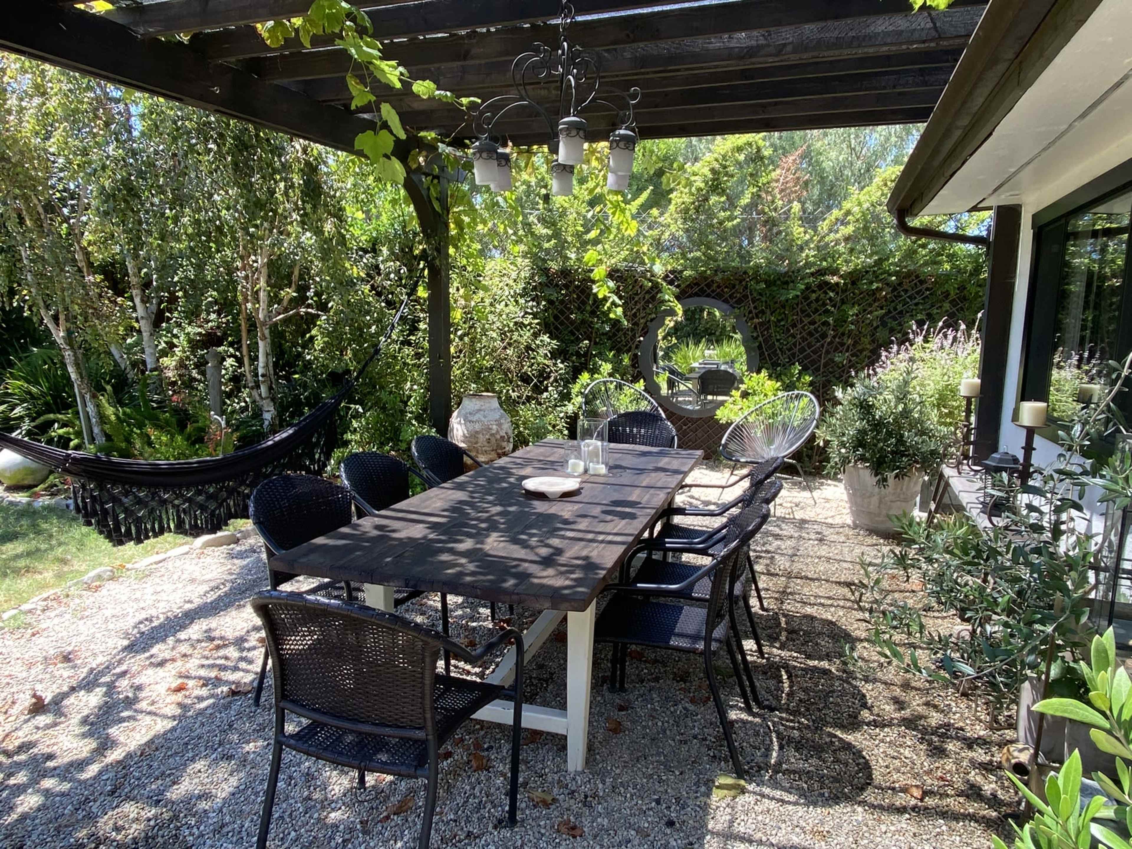 A long table surrounded by black chairs is set under a pergola adorned with greenery, next to a hammock and various plants.