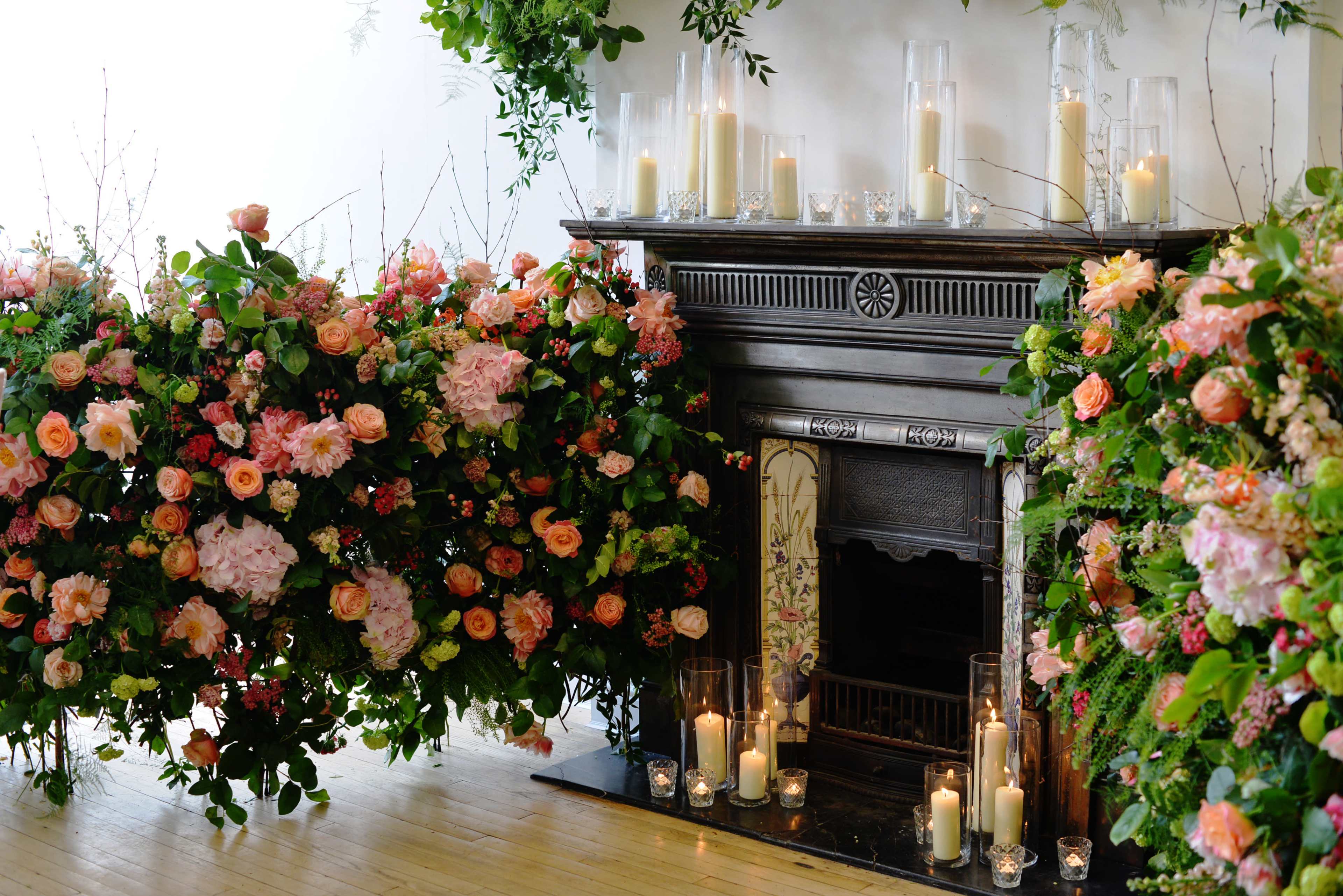 A fireplace is surrounded by large arrangements of pink and orange flowers, with candles placed on the mantel and in front of it.