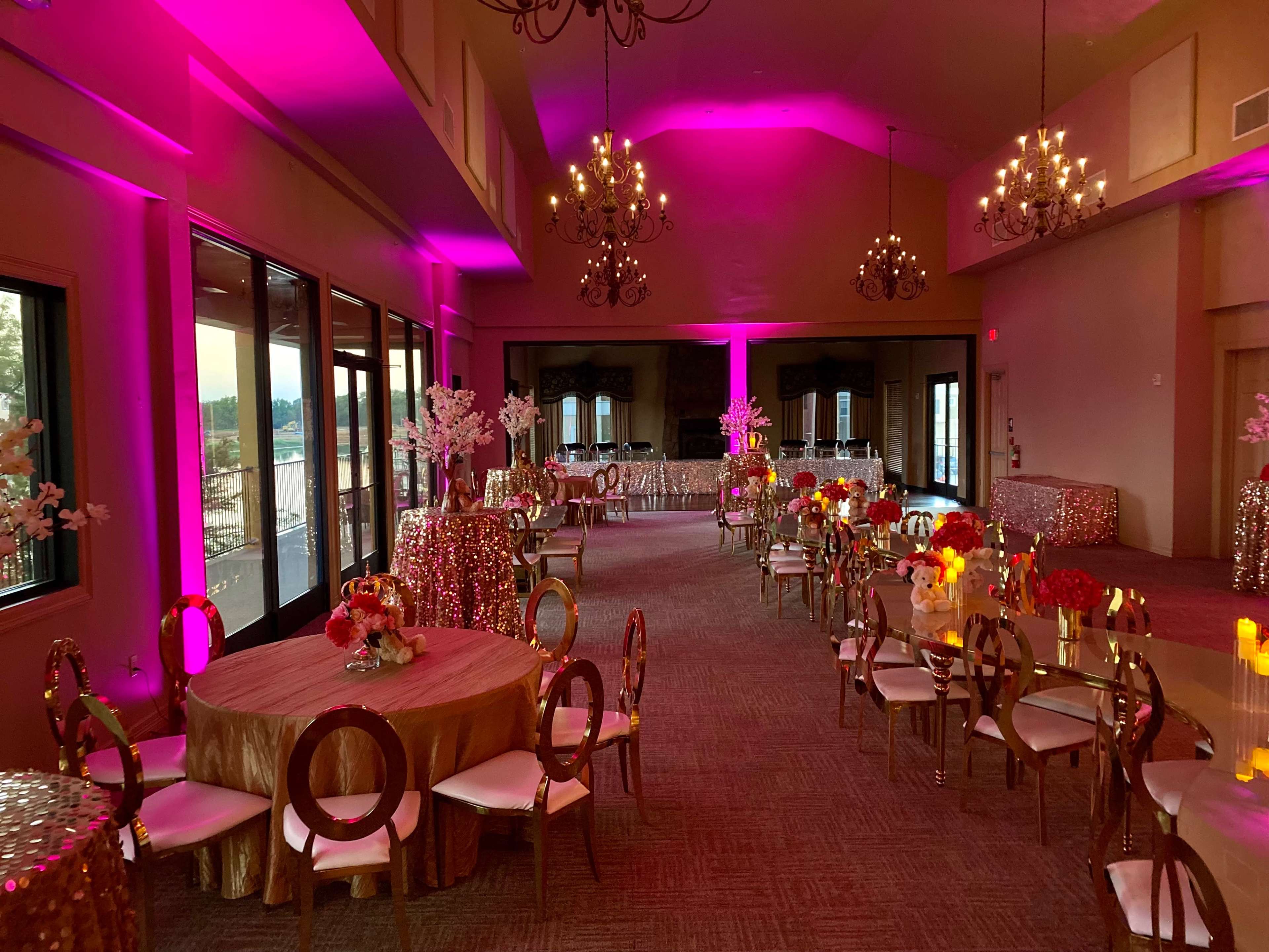 A decorated event space with pink lighting, elegant round tables, and floral centerpieces.