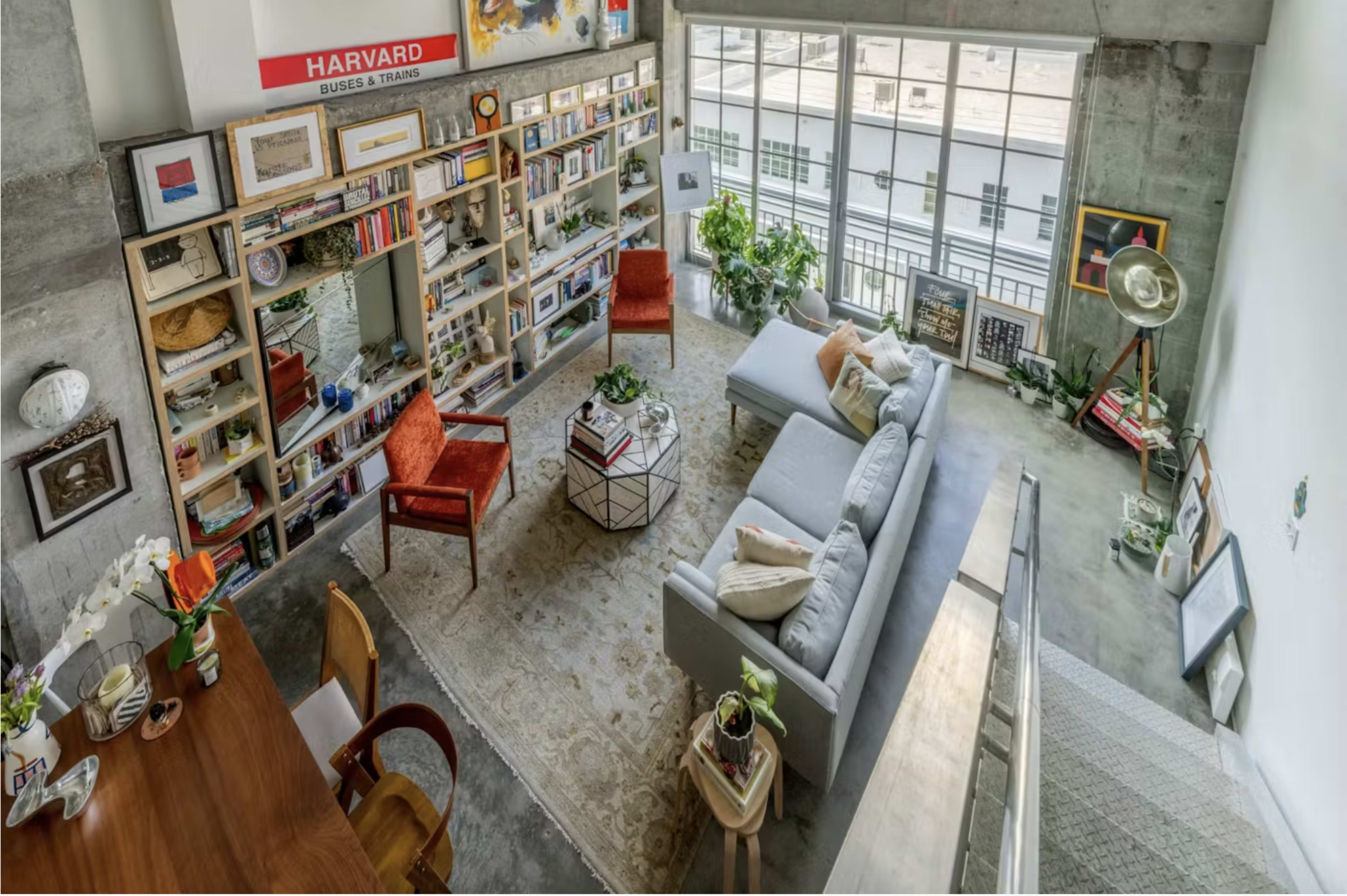 A modern living room with a gray sofa, a coffee table, and bookshelves filled with books and decorative items, surrounded by plants and natural light coming through large windows.