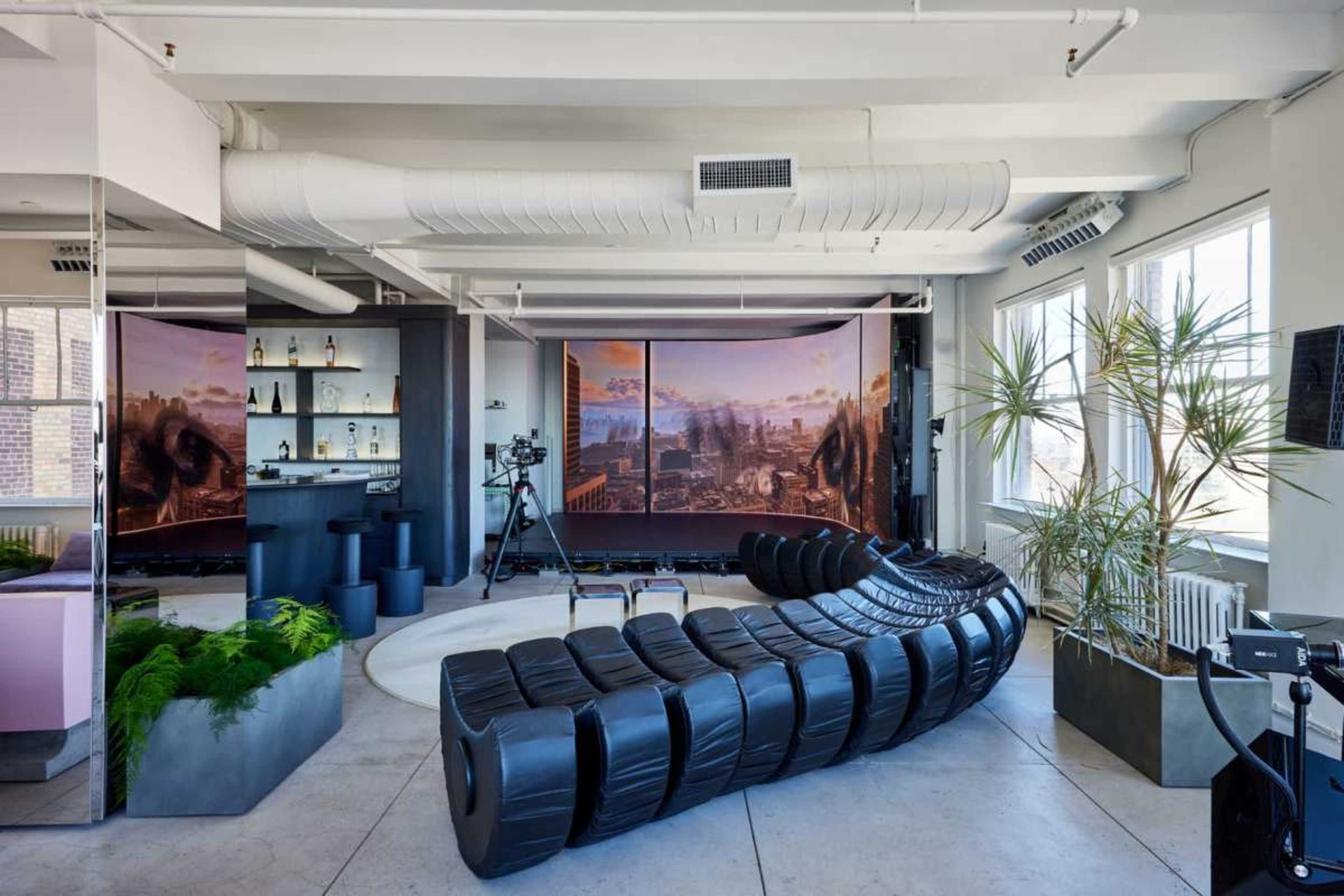 The image shows a modern open-concept living space featuring a curved black sofa, a bar area with bottles, a camera setup, and large windows allowing natural light to enter.