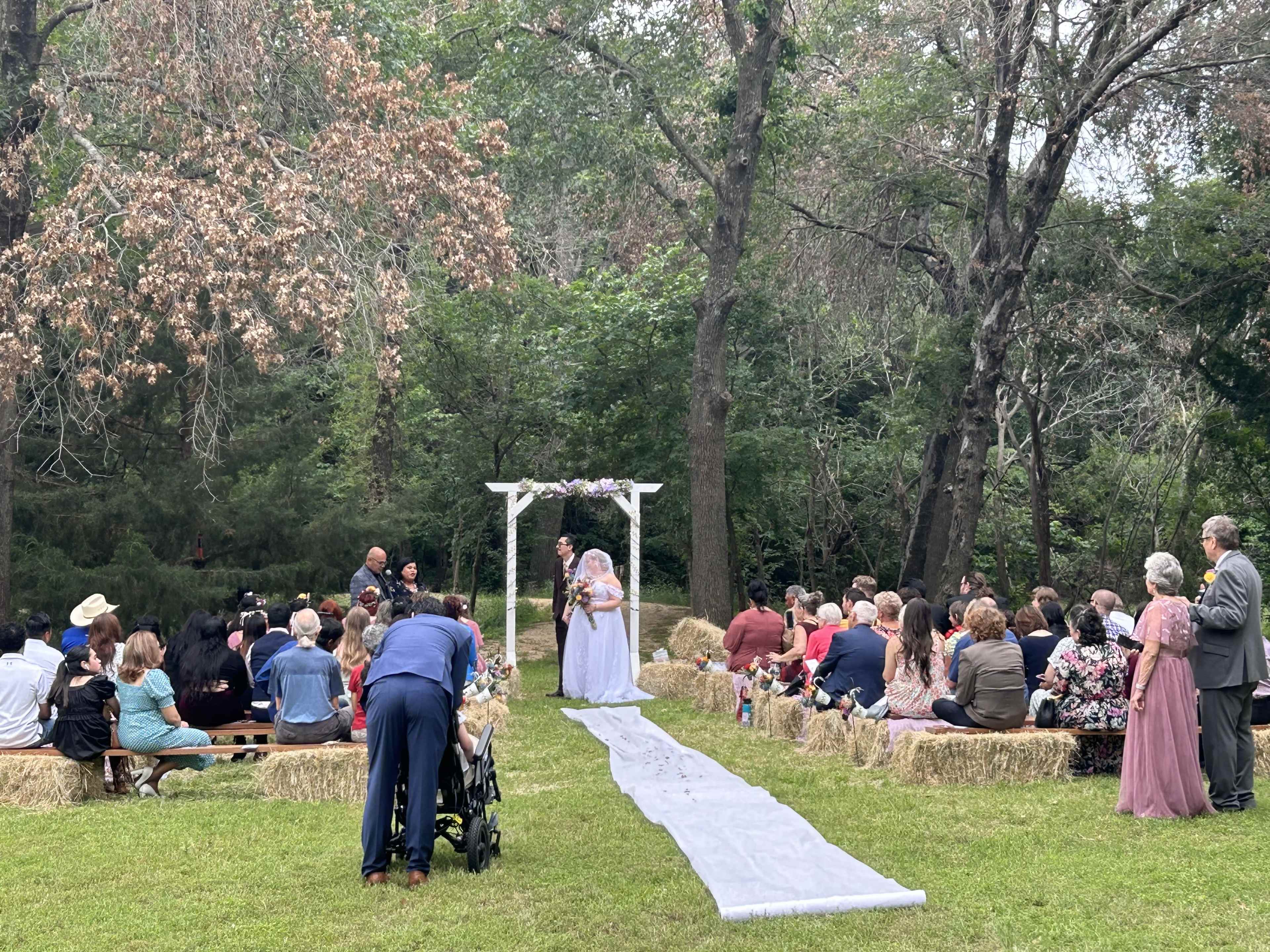 A wedding ceremony takes place outdoors under a floral arch, with guests seated on hay bales arranged on either side of a white aisle runner.
