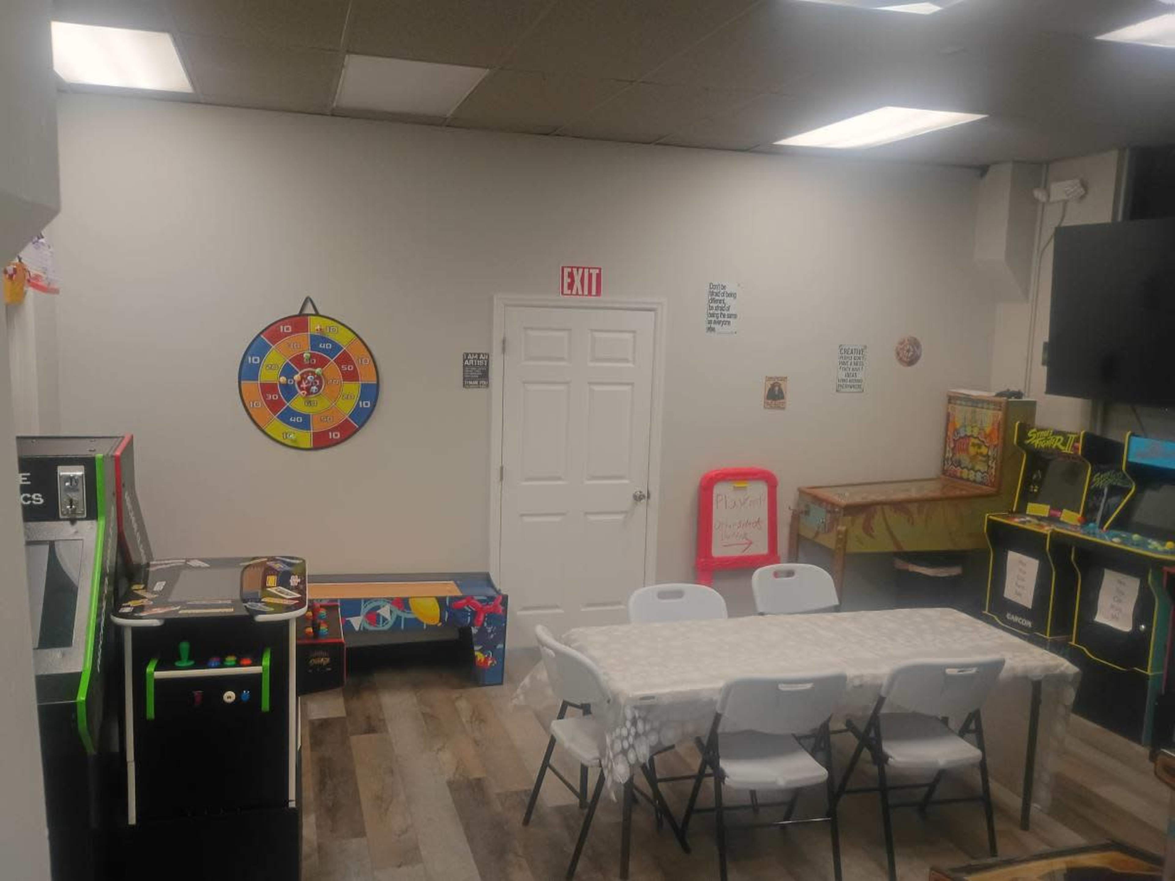 The image shows a small recreational room featuring an assortment of arcade machines, a dartboard on the wall, a table with chairs, and an exit sign.