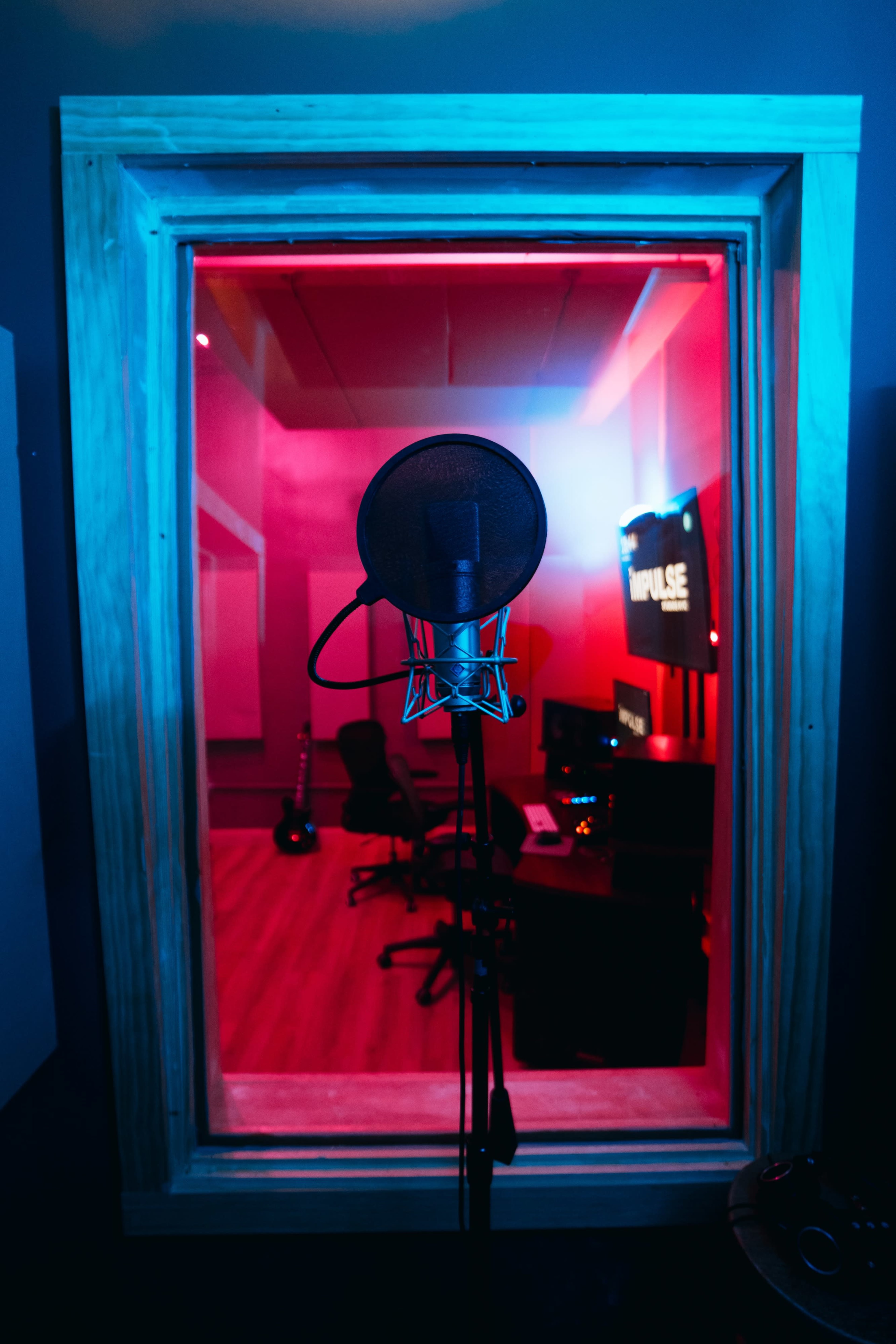 A microphone stands in a soundproof booth with colored lighting, overlooking a studio workspace equipped with a guitar and computer equipment.