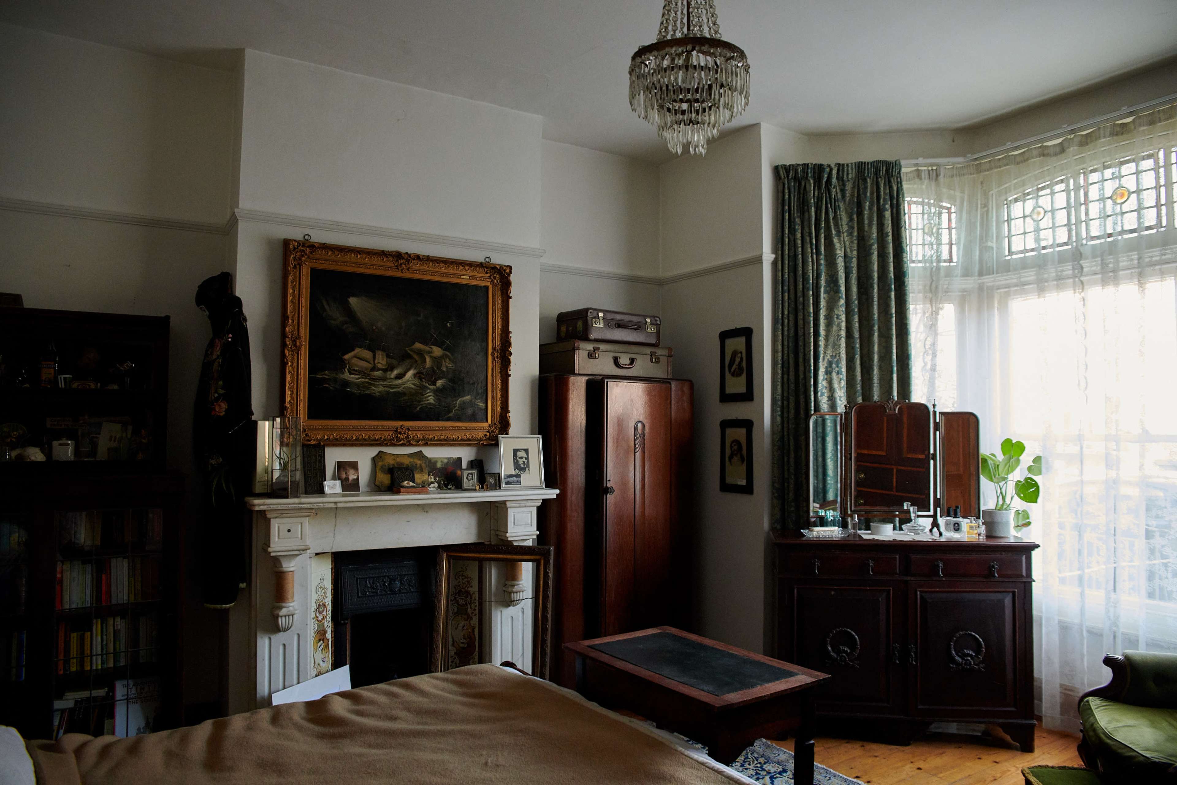 A vintage bedroom featuring a fireplace with a painting above it, an ornate wardrobe, a dresser with a mirror, and large windows with draped curtains.