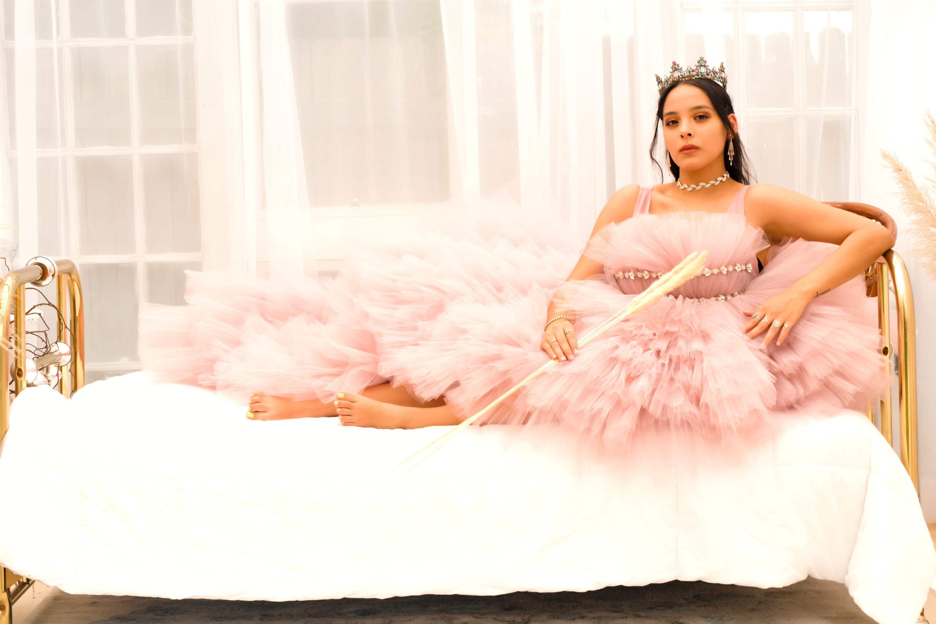 A woman in a pink tulle dress is seated on a bed, holding a decorative arrow while wearing a crown.