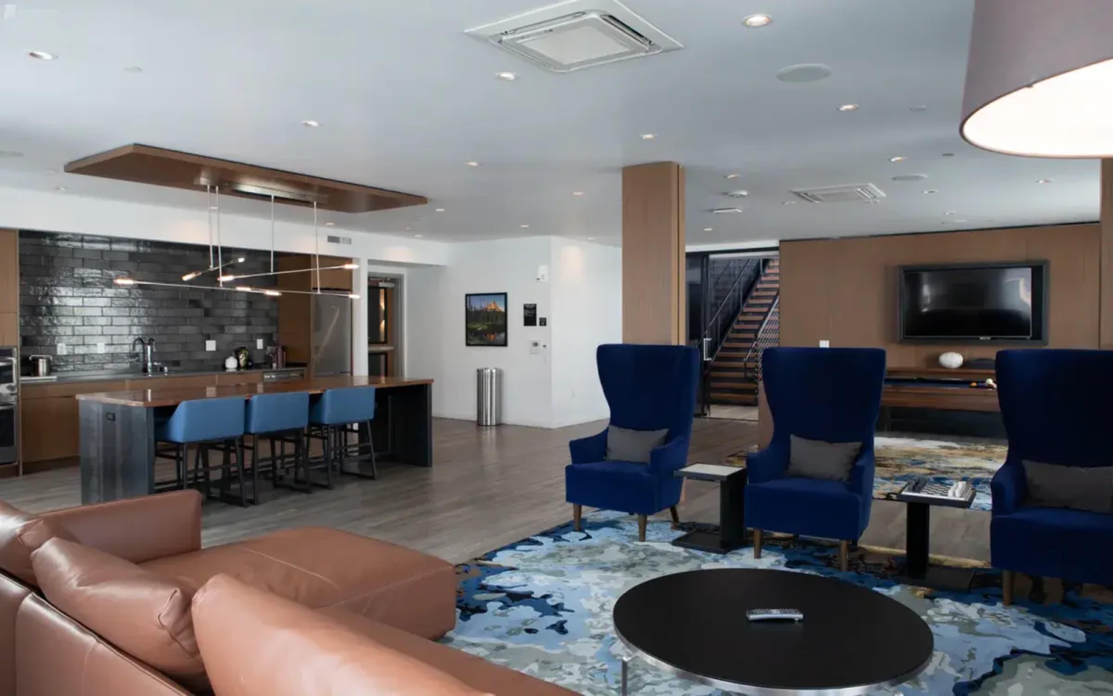 A modern open-concept living area featuring a kitchen with bar seating, two blue armchairs, a brown leather sofa, and a staircase in the background.