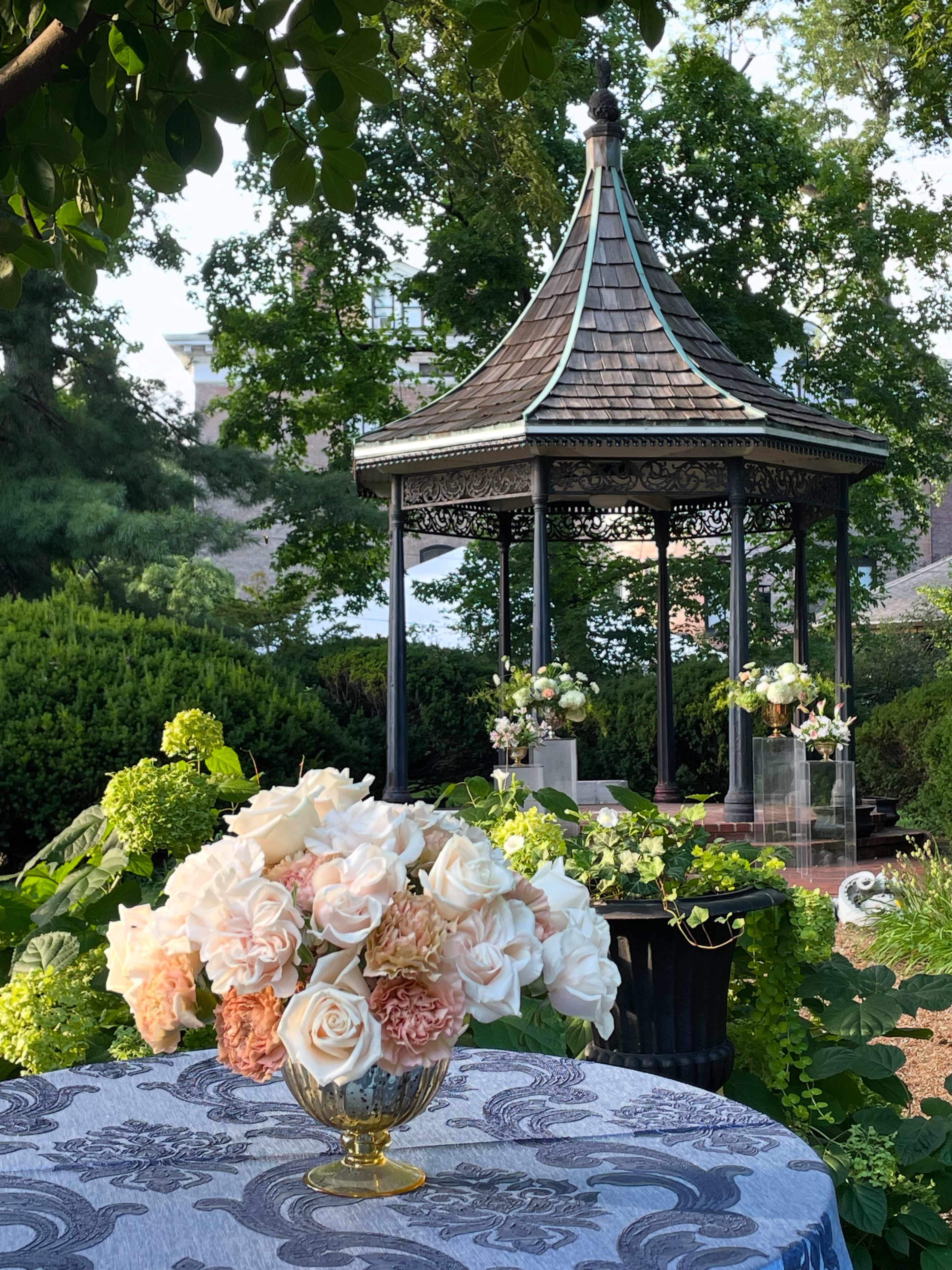 A round table with a floral arrangement sits in the foreground, while a gazebo surrounded by greenery is visible in the background.