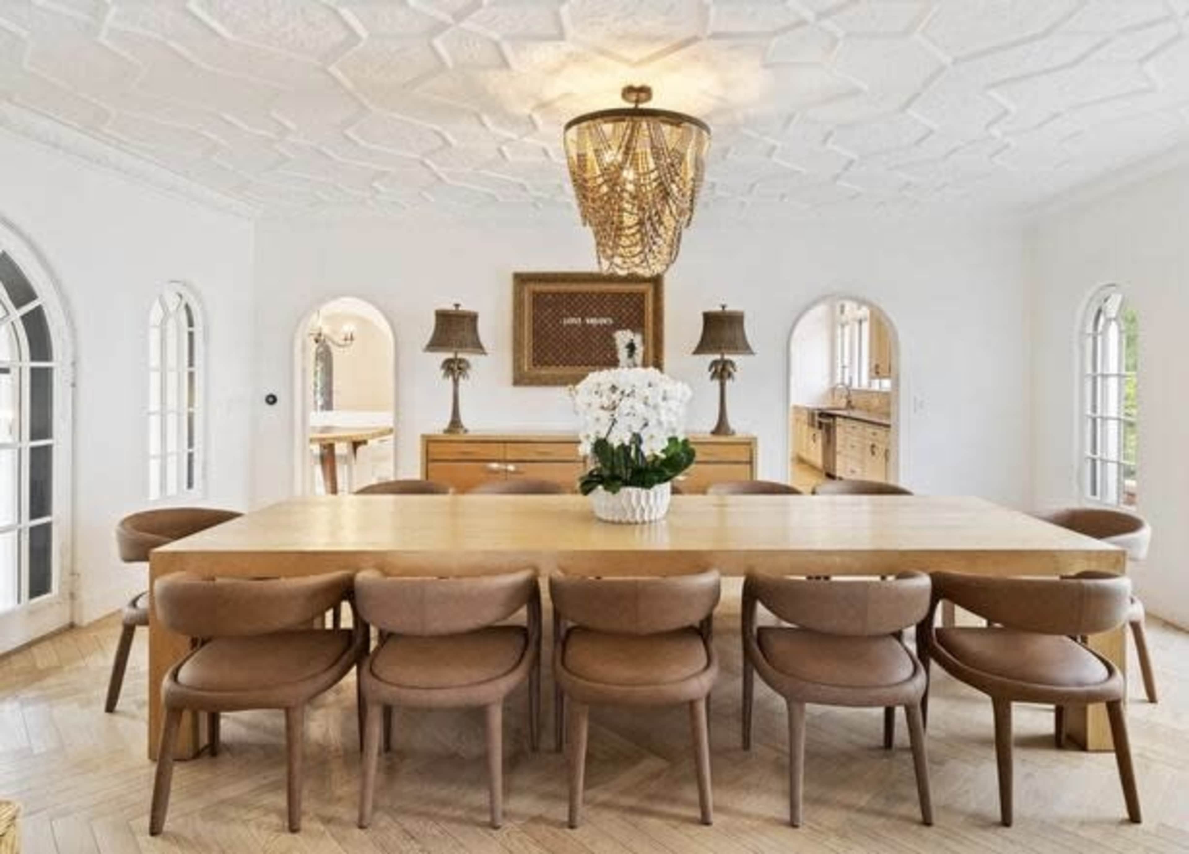 The image shows a spacious dining room featuring a large wooden table surrounded by ten upholstered chairs, with a chandelier overhead and decorative lamps in the background.