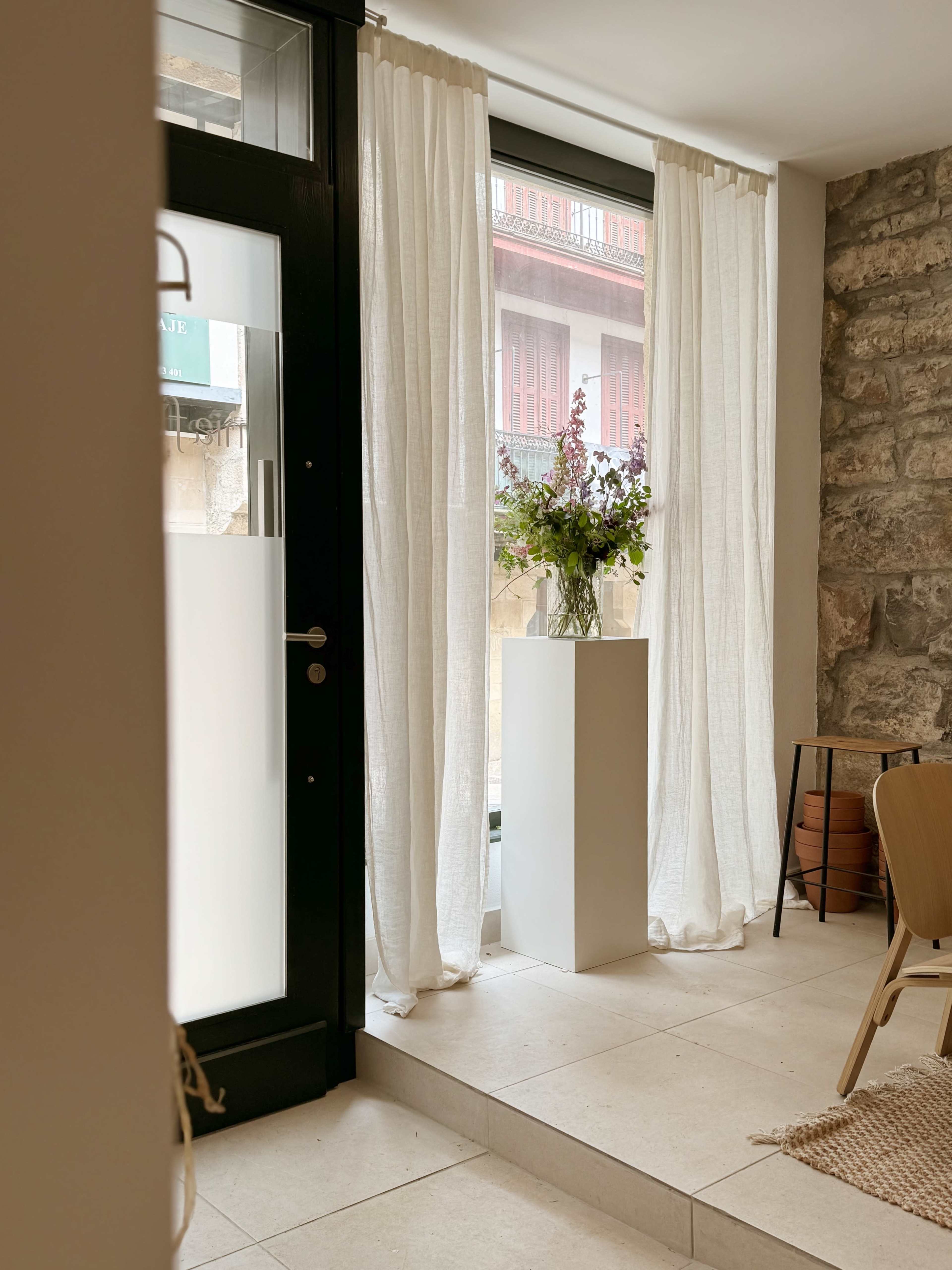The image shows a bright interior space with large windows dressed in sheer white curtains, a potted plant on a pedestal, and a wooden chair.