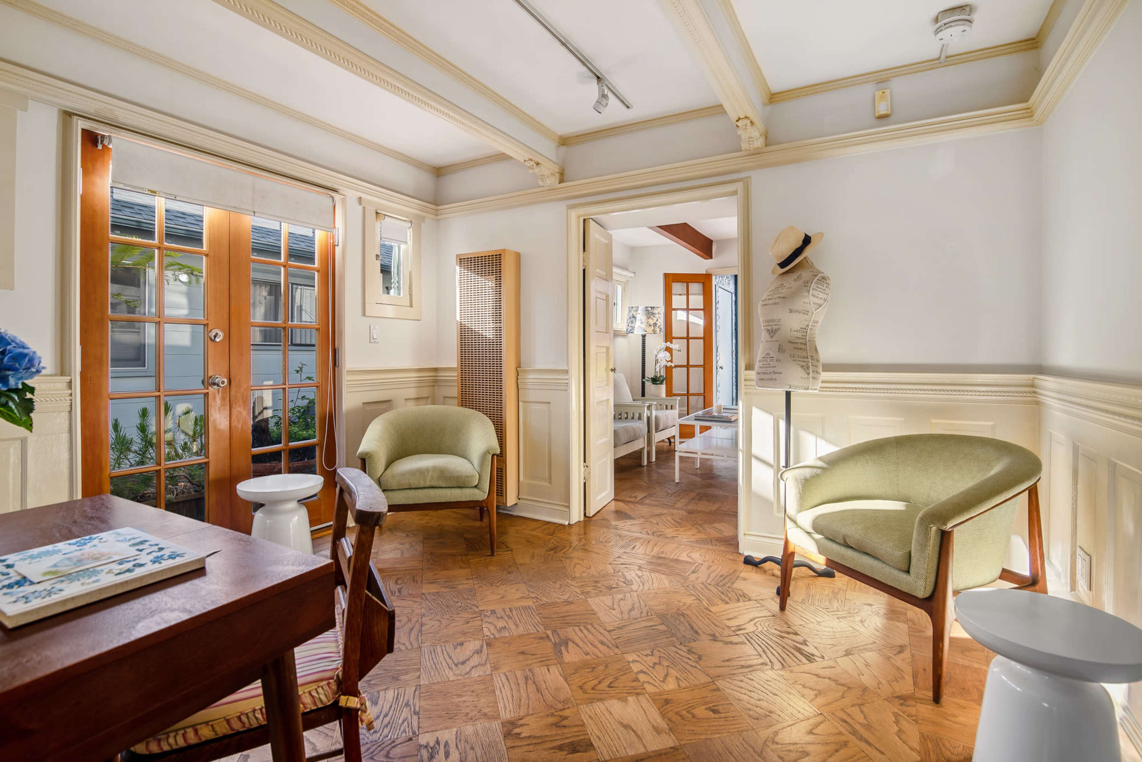 A cozy interior features a dining table, two green chairs, and a mannequin, with natural light streaming through glass doors and a doorway leading to another room.