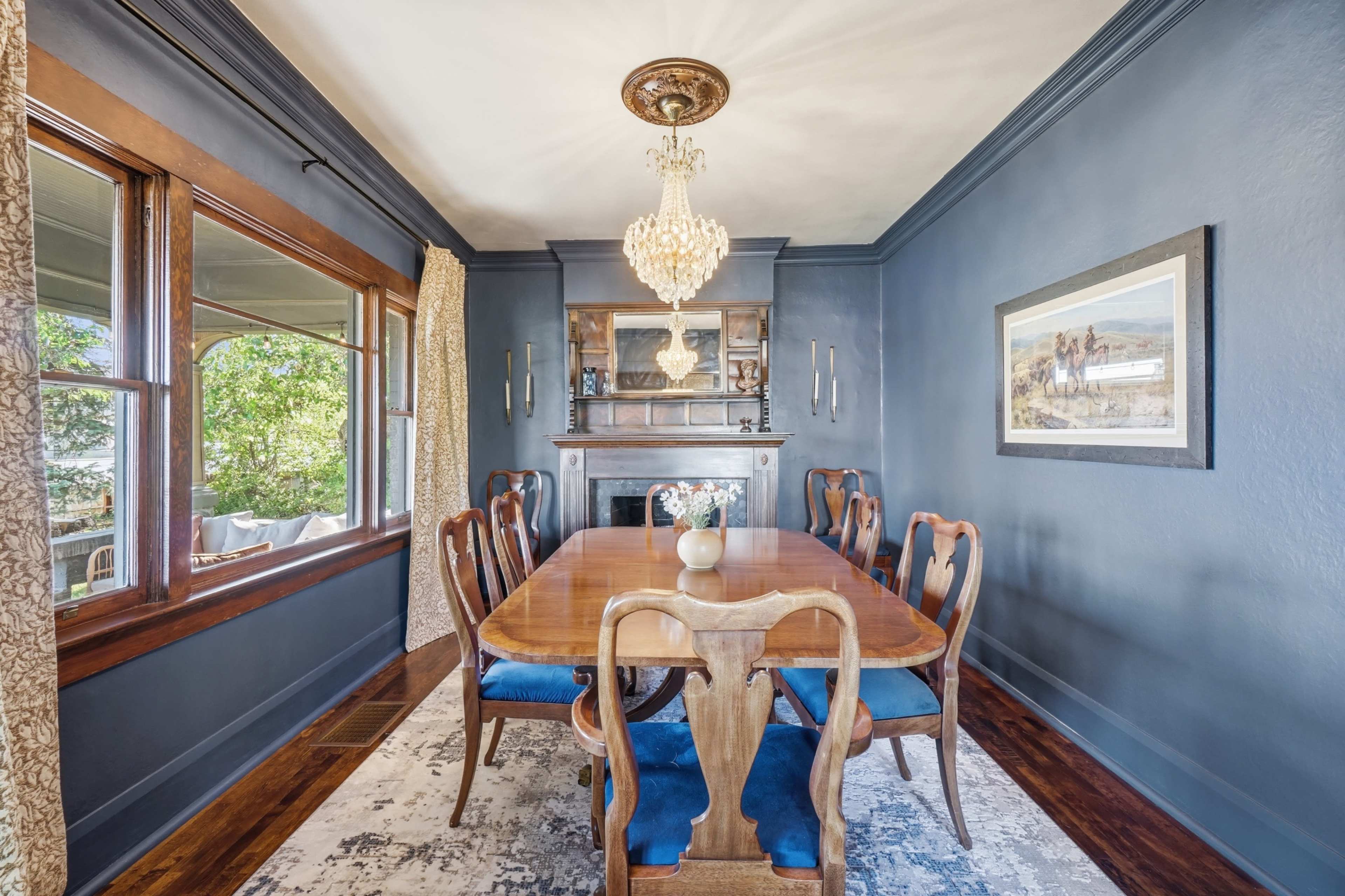 The image shows a dining room with a wooden table surrounded by eight chairs, a chandelier hanging from the ceiling, and large windows providing natural light.