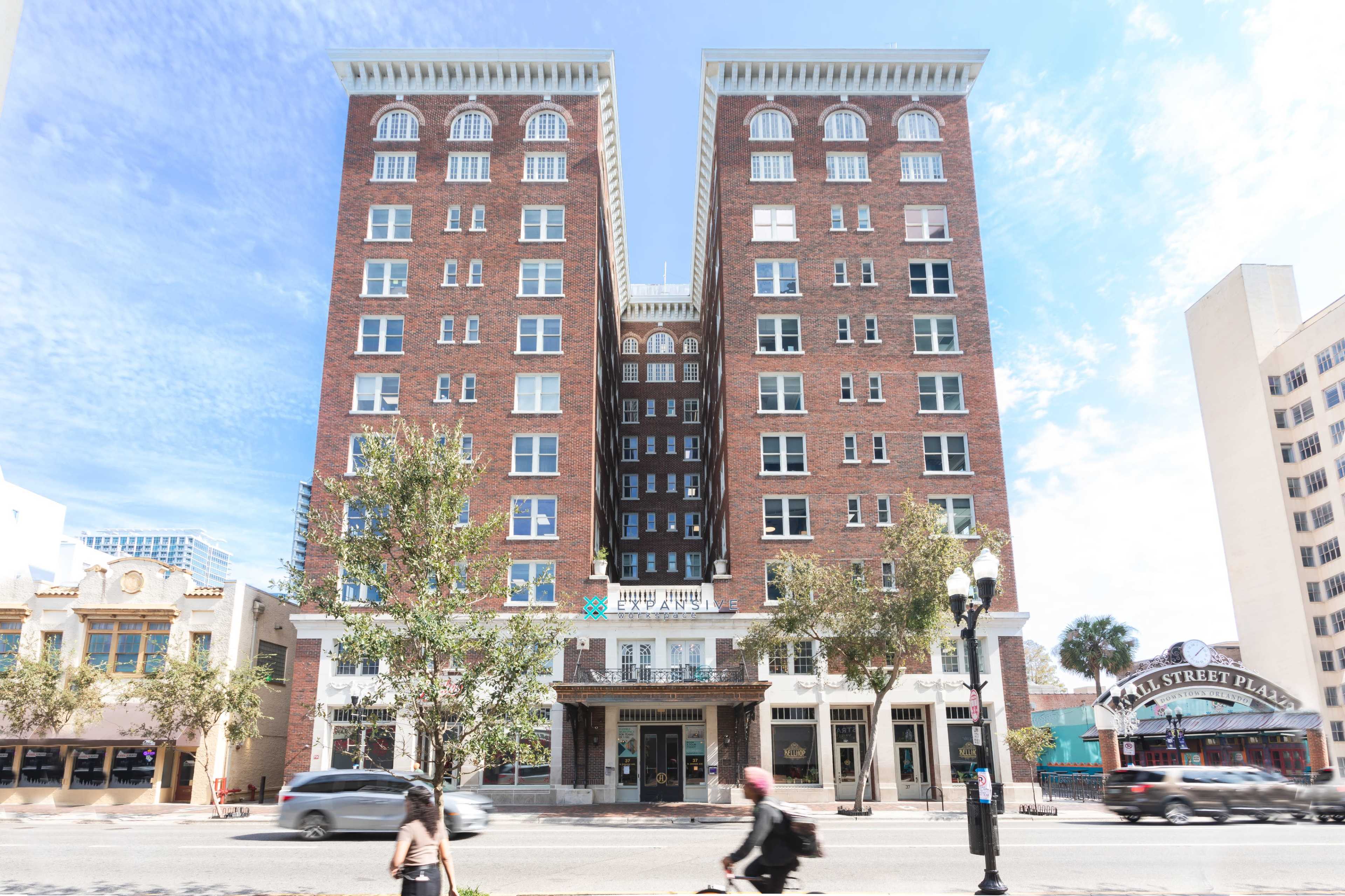 The image depicts a large brick building with several stories located on a city street, flanked by trees and a nearby sign for a shopping area.