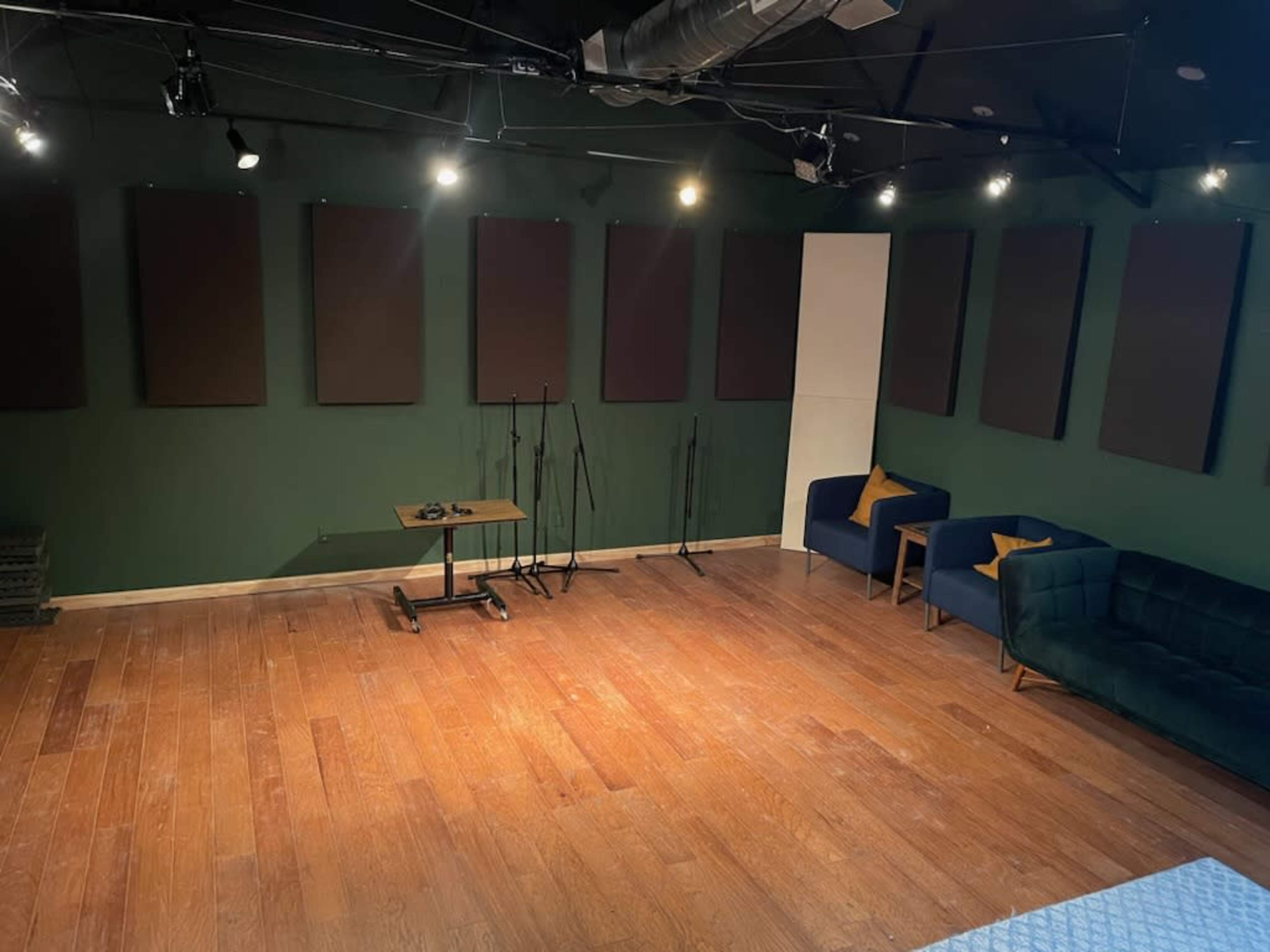 A music or recording studio with wooden flooring, green walls, multiple soundproof panels, and seating arrangements including two couches and a table.